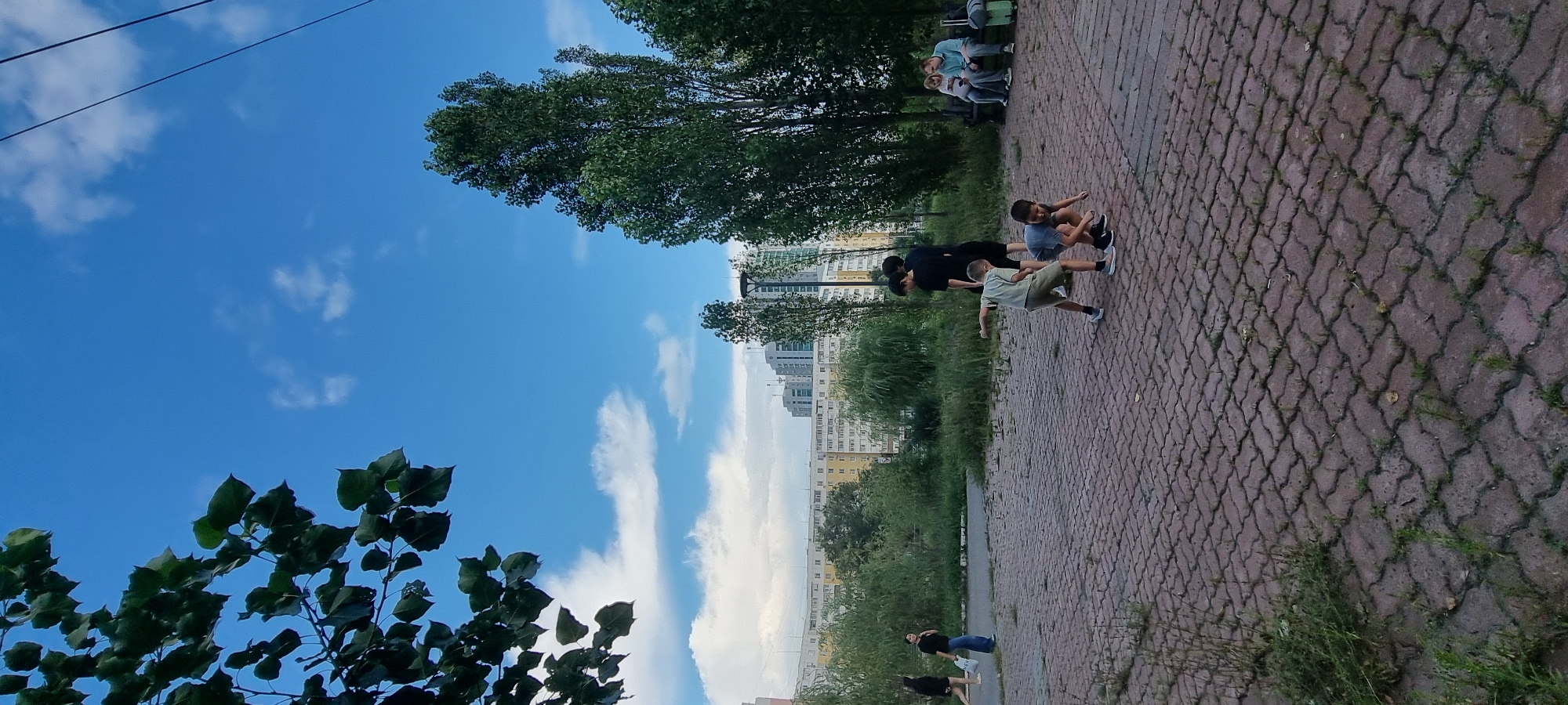 In Aktobe, Kazakhstan, on a cloudy day with a blue sky, a group of children are playing in a paved area near trees and apartment buildings. The children, appearing to be between 5 and 10 years old, are engaged in a game, with two of them in the foreground in a crouched or bending position. Further back, two other children are standing. Two adults are seated on a bench to the right, one looking at a phone. In the background, there are tall apartment buildings and lush green trees. A woman is walking away from the camera on the left side of the image. The ground is covered with interlocking brick pavers with some grass growing through the cracks. Overhead, power lines are visible against the sky.