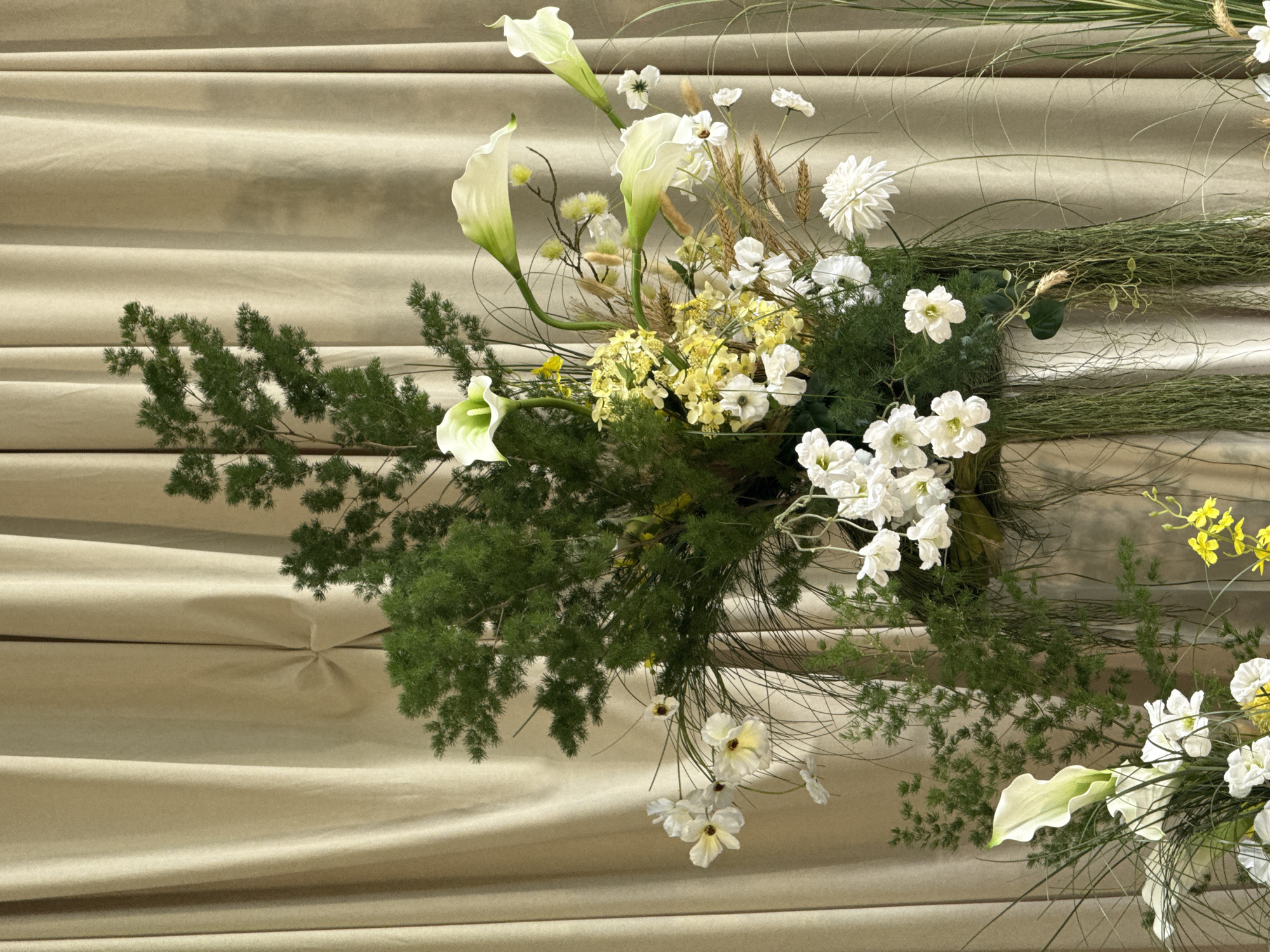 This image depicts a tall, elaborate floral arrangement situated indoors against a backdrop of draped, pleated beige curtains, located in Perm, Russia. The arrangement is composed of a prominent selection of natural and possibly artificial elements. Dominant features include dark green, feathery foliage, resembling Asparagus fern, forming a substantial part of the arrangement's height and volume. Interspersed within the green are several elegant white calla lilies, some exhibiting pale green throats, with gracefully curved stems. Numerous smaller white flowers with yellow or dark centers, resembling cosmos or anemones, are distributed throughout, along with denser clusters of layered white flowers akin to dahlias or chrysanthemums towards the bottom right. Accents of small yellow flowers, possibly solidago or hydrangeas, are visible, along with scattered brown, spiky wheat stalks and some fluffy yellow seed heads. Long, thin blades of grass-like foliage and cascading stringy green tendrils contribute to the arrangement's texture and form. The background curtains show vertical folds and a subtle seam with a small knot-like detail. No individuals, actions, or specific interactions are visible within the frame. The scene presents a static, decorative display.