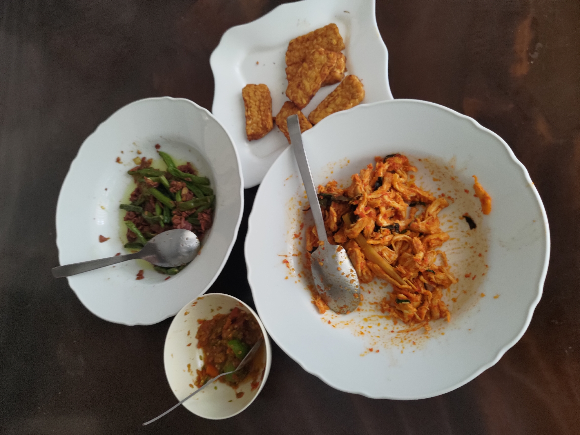 A bird's-eye view captures three white plates of partially consumed Indonesian food arranged on a dark brown wooden surface, possibly a table in a restaurant in Depok, Indonesia. The plate on the left contains a small portion of what appears to be sambal, a spicy chili paste, with a spoon resting inside. To its right, a larger plate holds a dish of green beans mixed with small pieces of meat or other ingredients, also with a spoon. In the foreground, a large white plate is filled with shredded chicken or meat in a reddish-orange spicy sauce, accompanied by a spoon and some of the sauce smeared on the plate's rim. To the right of this plate, a separate white square plate displays several pieces of fried tempeh, a fermented soybean cake, arranged in a pile. The lighting suggests an indoor setting, possibly in the afternoon.