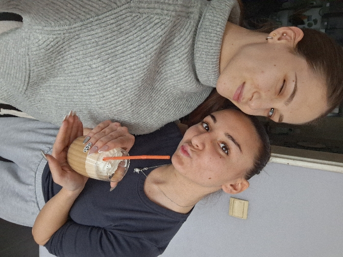 The image depicts two young adult females in a close-up, informal indoor setting. While the prompt specifies the location as Sofia, Bulgaria, the visual evidence within the image does not provide specific markers to pinpoint a precise location within that city.
The subject on the left, positioned slightly forward, has brown hair and is wearing a light grey, ribbed turtleneck sweater. She is winking with her right eye and displaying a subtle smile. A small gold stud earring is visible in her left earlobe, along with a small gold hoop earring positioned slightly lower. The subject on the right, positioned slightly behind and to the right, has her brown hair pulled back from her face. She is wearing a dark grey, long-sleeved t-shirt and light grey sweatpants. Her lips are pursed in a pout. She is holding a clear glass containing a layered beverage with a light brown liquid base, topped with white whipped cream and an orange plastic straw. Her fingernails exhibit a decorative design, with some nails featuring white tips and others displaying a black and white animal print pattern. A delicate silver chain necklace with a small charm is visible around her neck. Her right hand cups the bottom of the glass while her left hand grips the upper portion. The background consists of a plain, light grey wall, with a small, light brown rectangular object, possibly a light switch or outlet cover, mounted on it to the right. A dark, indistinct area is visible to the far left, suggesting a doorway or furniture. The interaction appears casual, indicative of a shared moment, possibly a selfie.