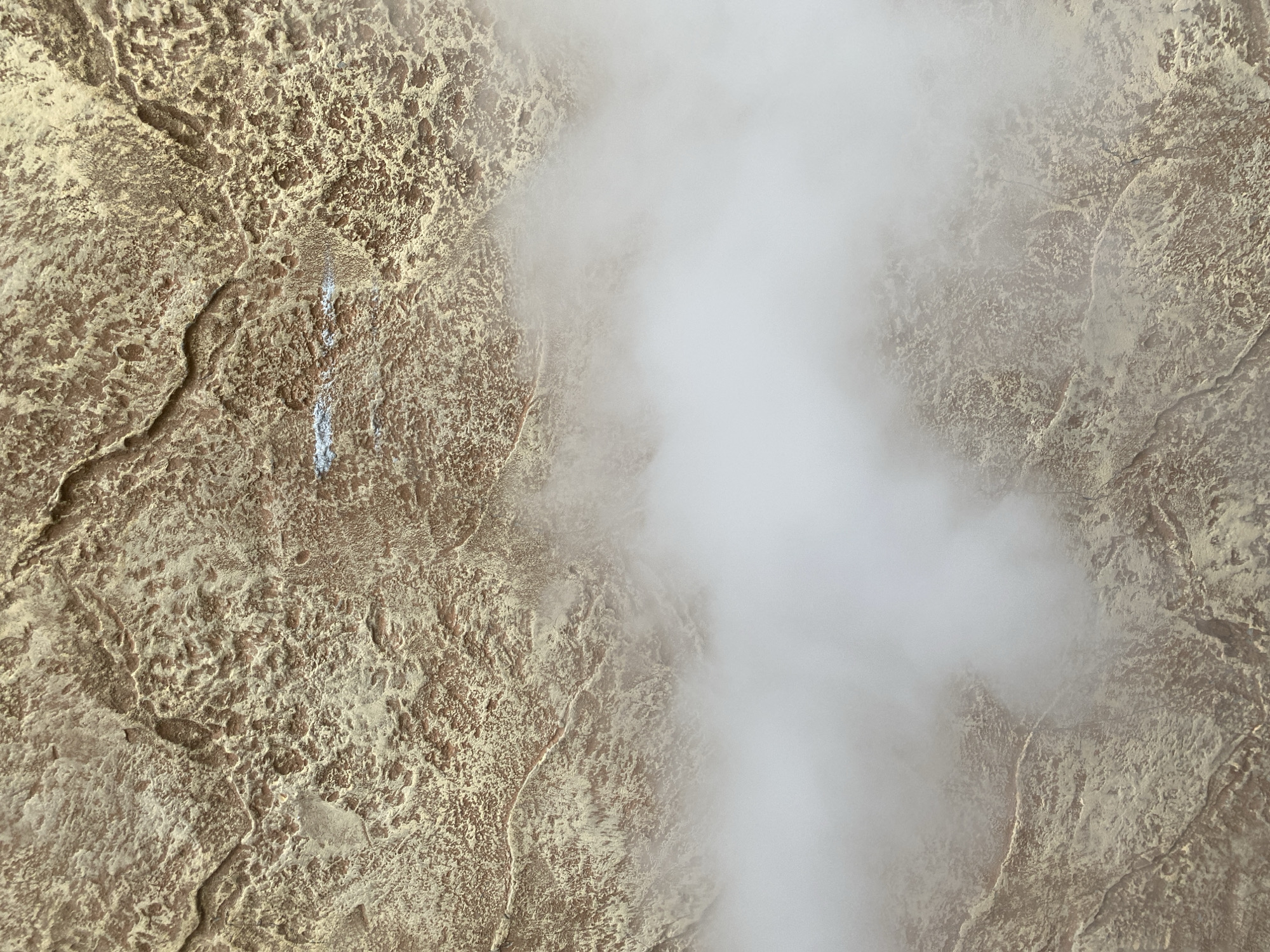 This image presents an aerial or close-up overhead view of a textured, rugged terrain, primarily composed of earthy brown and yellowish-sandy tones. The surface features intricate patterns of cracks, fissures, and varying elevations, resembling weathered rock or mineral deposits. A significant portion of the right side of the frame is dominated by a large, dense plume of white mist or steam, which appears to be actively rising from or across the terrain, partially obscuring the ground below. In the upper-left quadrant of the textured surface, a distinct, irregular streak of light blue or white material is visible. The scene suggests a natural geological phenomenon, such as a hot spring, geyser, or fumarole, characterized by steam emission. No people, man-made objects, or visible text are present. The lighting is diffuse, and the time of day or specific weather conditions, other than the mist, are not discernible. The location context is Troyan, Bulgaria.