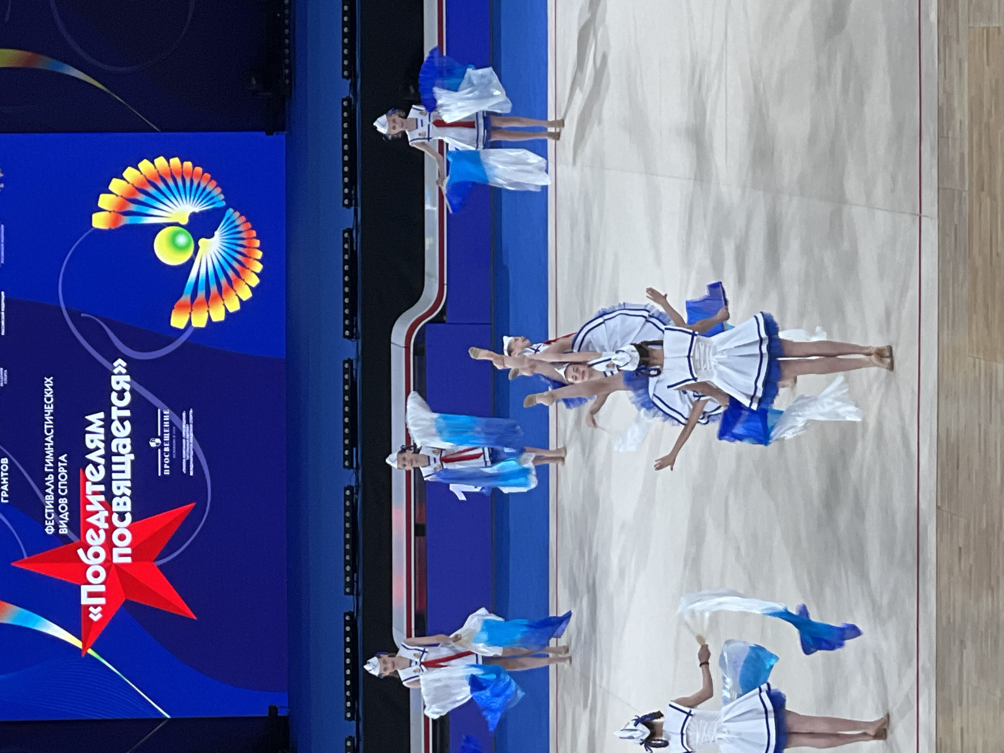 The image depicts a synchronized gymnastics performance. Several young female athletes, dressed in white and blue sailor-inspired uniforms with matching caps, are performing on a large, light-colored mat. They are holding and manipulating blue and white fabric streamers, creating flowing patterns. The athletes are in various poses, some standing, some bending, and at least two are in mid-air with legs extended. The background features a large blue screen displaying text in Russian, including "ГРАНТОВ" and "ФЕСТИВАЛЬ ГИМНАСТИЧЕСКИХ ВИДОВ СПОРТА «ПОБЕДИТЕЛЯМ ПОСВЯЩАЕТСЯ»," which translates to "GRANTORS" and "FESTIVAL OF GYMNASTICS SPORTS 'DEDICATED TO THE WINNERS'." A stylized graphic of a colorful fan-like object with a green sphere is also visible on the screen. The overall environment suggests an indoor arena or performance hall. The specific location in Moscow, Russia, cannot be determined from the visual information provided in the image.