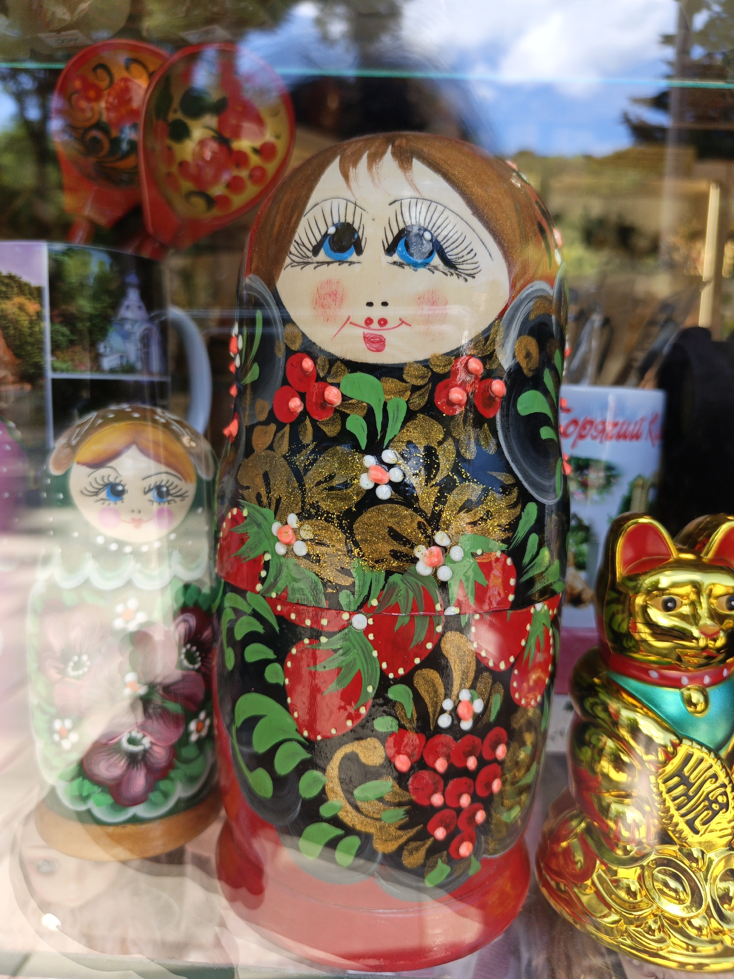 This close-up shot captures a vibrant display of decorative items, likely viewed through a shop window or display case in Goryachiy Klyuch, Russia. In the foreground, two hand-painted matryoshka dolls (Russian nesting dolls) are prominent, visible behind a reflective glass surface. The largest doll, centrally positioned, features a bright red base and a black bodice intricately decorated with gold glitter, red berries, green leaves, and white flowers. It has a cheerful face with striking blue eyes, long lashes, and rosy cheeks.
To the left, a smaller matryoshka doll is partially obscured, adorned with a light green design featuring purple flowers and white accents, and a similarly smiling face with blue eyes. On the far right, a shiny golden Maneki-neko (waving cat) figurine is visible, adding an eclectic element to the display.
The reflective glass surface reveals several background elements and reflections. In the upper left, two traditional red wooden spoons, decorated in a Khokhloma style with floral patterns, are reflected. Behind the dolls, a white mug is partially visible, bearing the Russian text "Горячий Ключ" (Goryachiy Klyuch), which is the provided location context. Reflections of a bright blue sky, green foliage, and a distant white building (possibly a church or chapel) suggest a sunny outdoor setting. The dolls' smiling faces contribute to a cheerful and inviting atmosphere for this souvenir display.