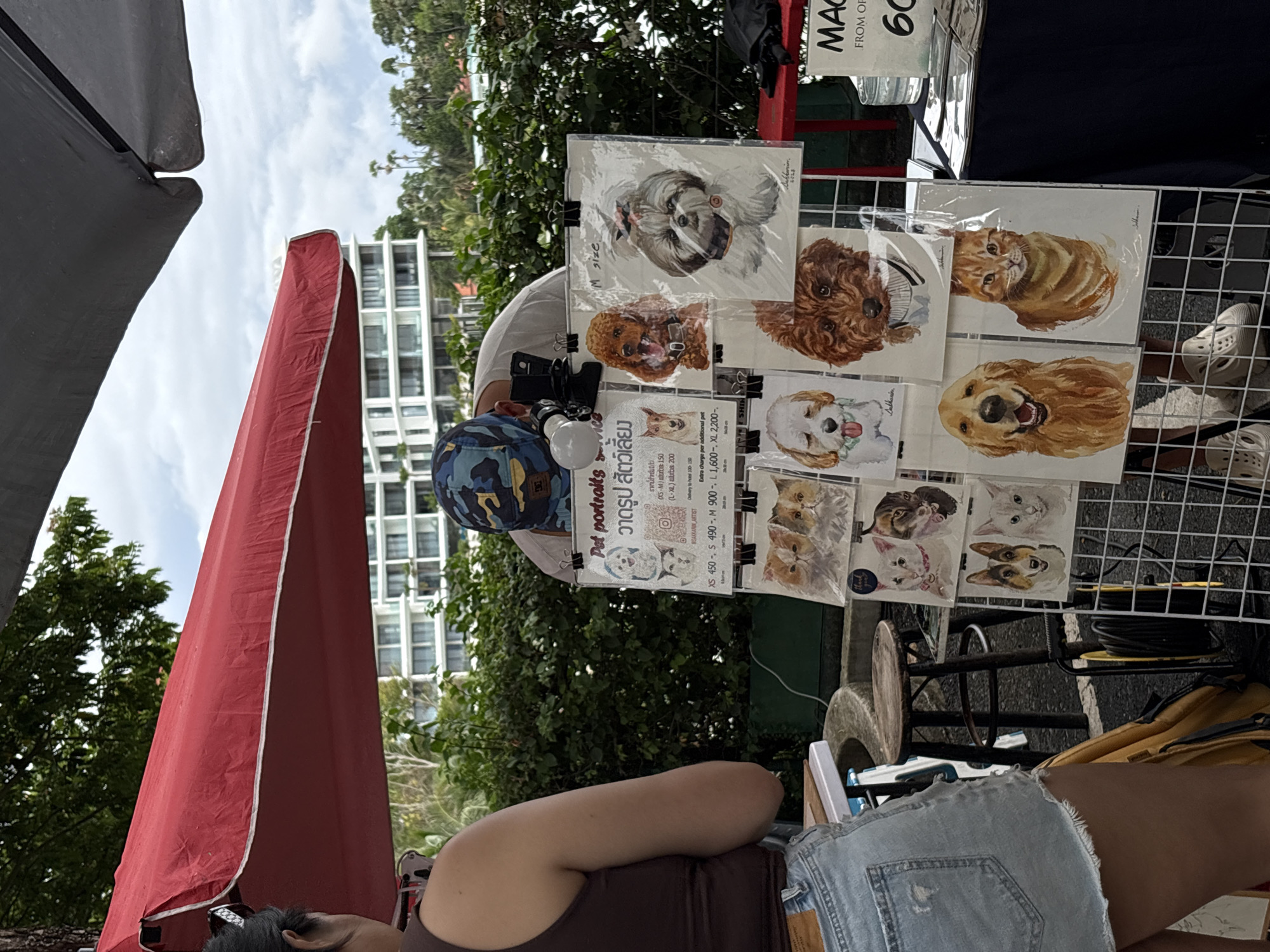 The image shows an outdoor stall displaying pet portraits in what appears to be a commercial area of Choeng Thale, Thailand. A woman, wearing a brown tank top and denim shorts, stands to the left, facing away from the camera. A man, wearing a camouflage baseball cap and a light-colored t-shirt, is bent over the stall, interacting with the displayed artwork. The stall features a grid display holding numerous watercolor pet portraits, primarily of dogs and cats, with some framed and others in protective sleeves. A sign in Thai and English advertises "Pet portraits" with prices listed in Thai Baht. Behind the stall, lush green foliage and the exterior of a modern, multi-story building are visible, suggesting a resort or hotel setting. The overall atmosphere is that of a marketplace or street fair, likely in a tourist-oriented location.