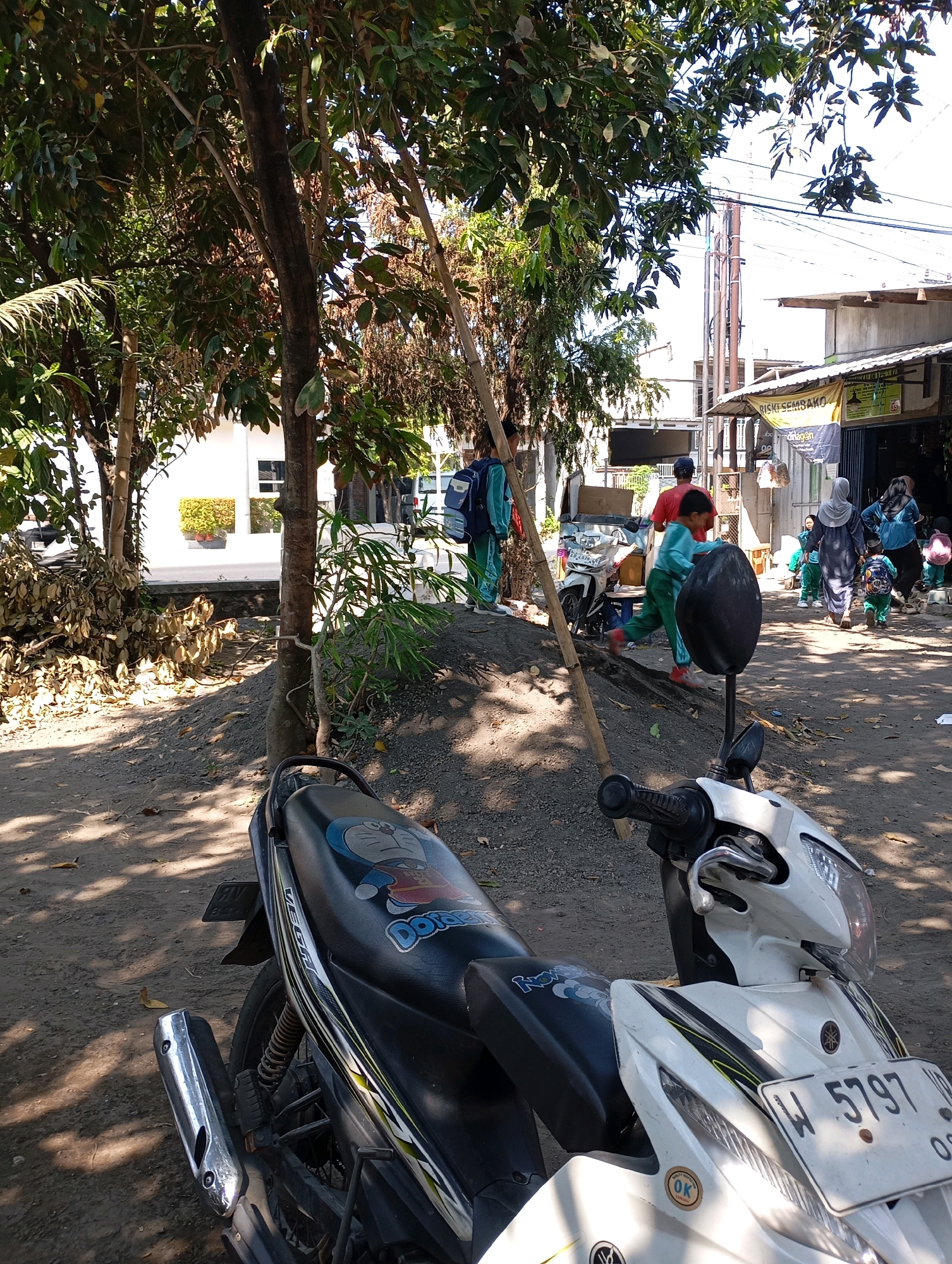 In Ketegan, Indonesia, a motorcycle is parked in the foreground, facing right. The motorcycle seat features a cartoon character, possibly Doraemon, and the license plate reads "W 5797". Behind the motorcycle, a dirt path slopes upwards towards a street. Several people, including children in green uniforms, are walking along the street. Some adults are also present, including a woman in a headscarf. Buildings line the street, with one appearing to be a shop with a banner that reads "RISKI SEMBAKO". Trees with green and dry leaves are visible on the left side of the image, with sunlight casting shadows on the ground. A wooden pole is propped against a tree.