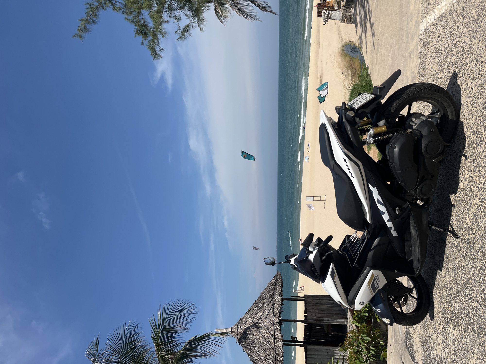 A white and black Yamaha NMAX scooter is parked on a textured concrete surface in the foreground. Behind the scooter, a sandy beach stretches to the turquoise ocean under a bright blue sky with scattered clouds. A thatched-roof hut with wooden supports stands to the left of the frame, partially obscured by palm fronds. Several kites are visible in the sky and on the beach, suggesting kite-surfing activities. Two small figures are present on the beach. The scene appears to be a coastal area in Mũi Né, Vietnam, a popular destination for water sports.