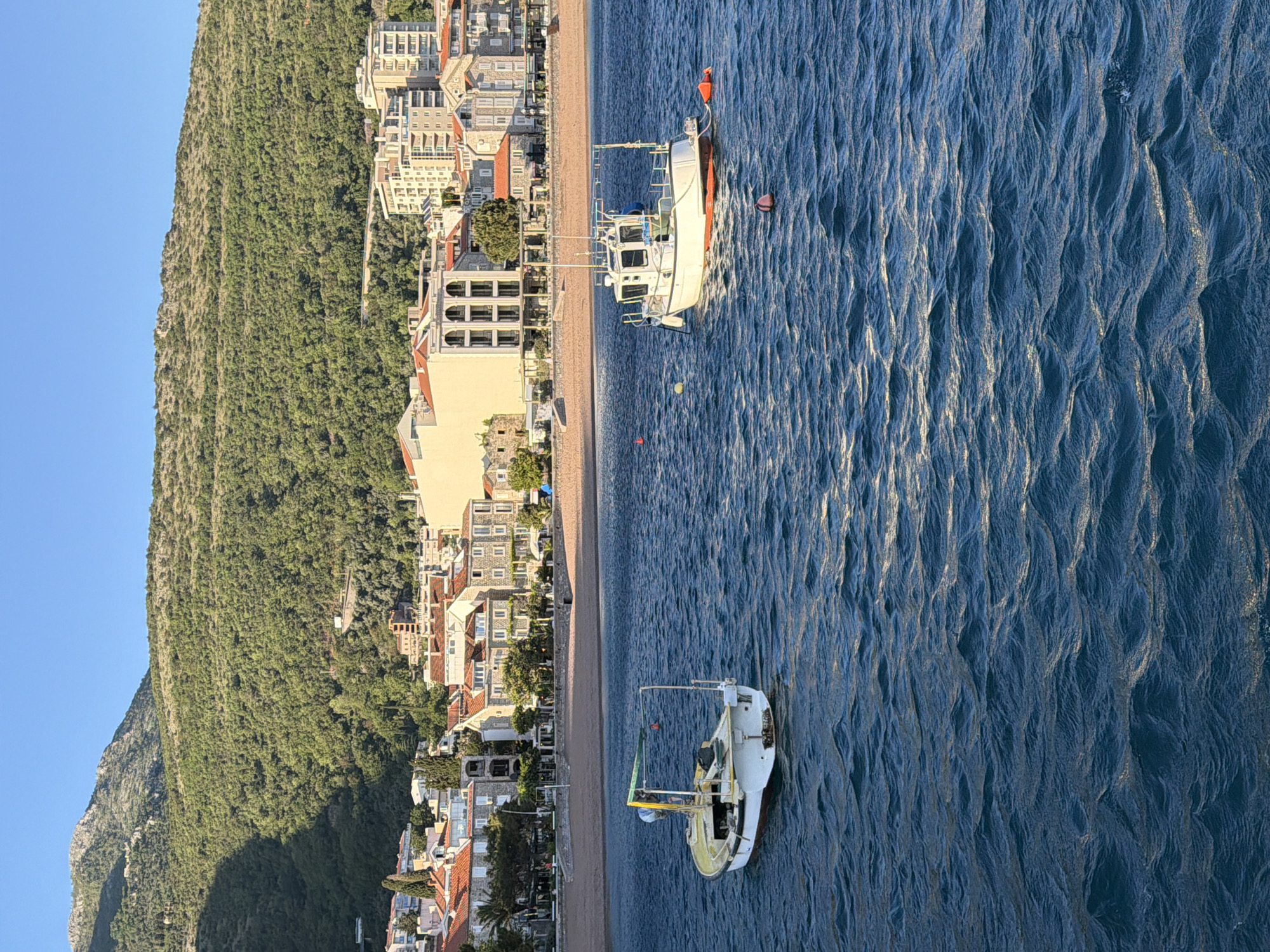 This media file presents a vibrant coastal landscape in Petrovac, Montenegro, captured from an elevated perspective. A sprawling, heavily forested mountain dominates the left side of the frame, its verdant slopes extending towards a built-up area.
Along the base of the mountain and bordering a sandy beach, numerous buildings form a town or settlement. Many of these structures are multi-story, exhibiting various architectural styles, and several are topped with distinctive terracotta roofs, common in Mediterranean regions. The sandy beach itself acts as a natural separator between the town and the sea.
The right side of the image features the deep blue waters of the sea, characterized by numerous small ripples that reflect the sunlight, creating shimmering patterns across the surface. Two boats are anchored in the water: a larger, predominantly white vessel positioned further out, and a smaller, white and yellow boat closer to the shore. Several buoys, including red and yellow ones, are scattered in the water.
The clear, bright blue sky suggests fair weather, and the soft, directional lighting illuminating the scene indicates either early morning or late afternoon. The overall impression is one of serenity and picturesque coastal beauty. No people are visibly discernible in the image, and there is no legible text on any of the boats or buildings.
