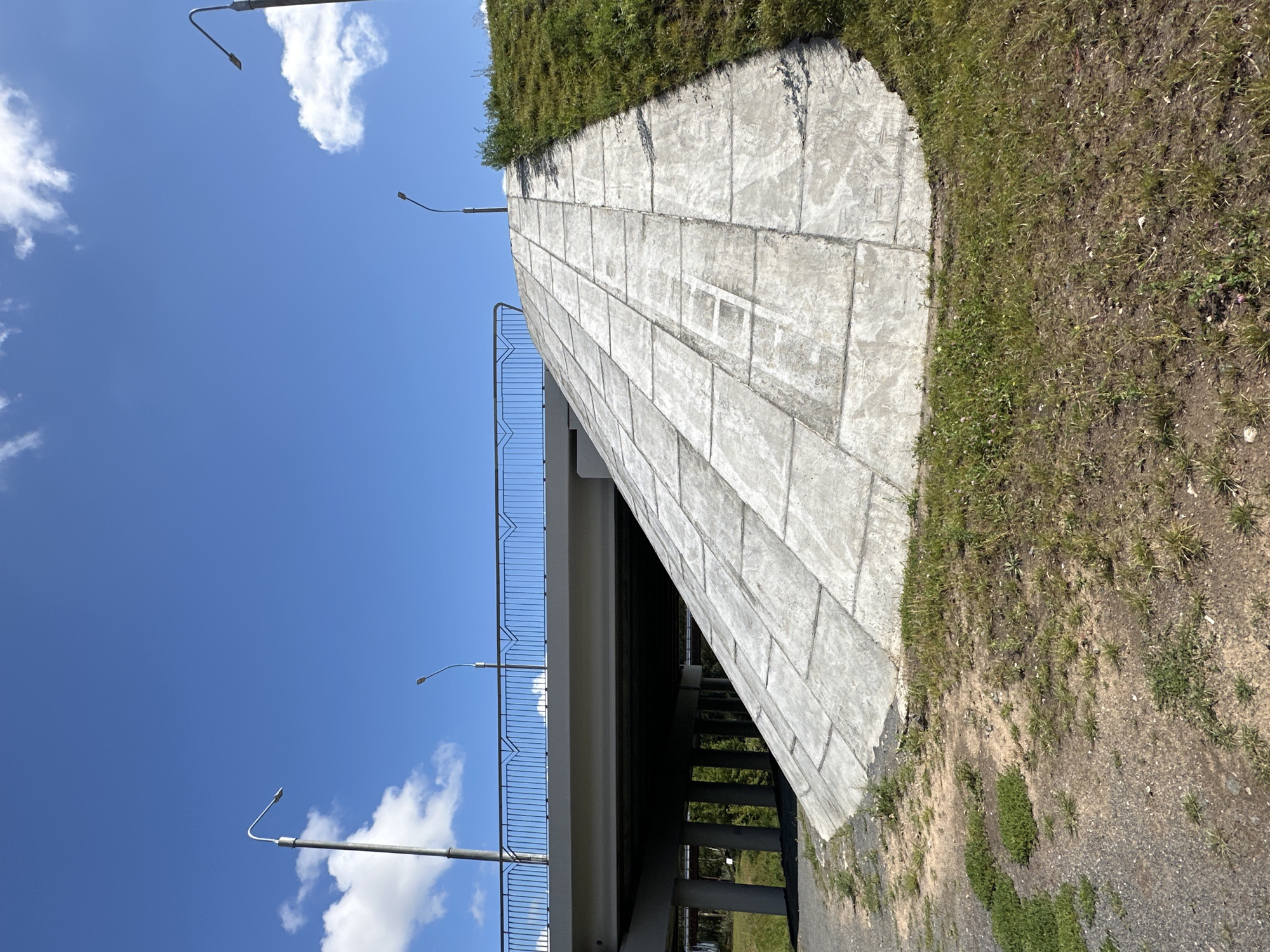 The image depicts a concrete ramp leading up to a bridge in Minsk, Belarus. The ramp is constructed from large, square concrete slabs and is flanked by a grassy embankment. A modern bridge with a metal railing spans across the scene, supported by concrete pillars. Two lampposts are visible along the bridge, and another is positioned on the grassy incline. The sky is a clear blue with a few scattered white clouds. The ground in the foreground consists of gravel and sparse vegetation. There are no people or vehicles present in the image.