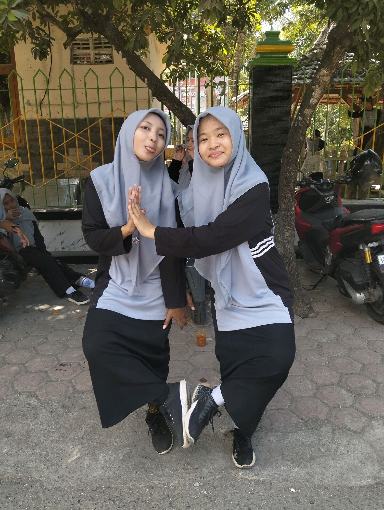 Two young women, wearing light gray hijabs and black long-sleeved shirts and skirts, are posing for a photo in an outdoor environment. The woman on the left is sticking her tongue out playfully, while the woman on the right is smiling at the camera and high-fiving the other woman. They are both wearing black sneakers. In the background, there is a fence, a tree, and a motorcycle. The scene appears to be in Paciran, Indonesia.