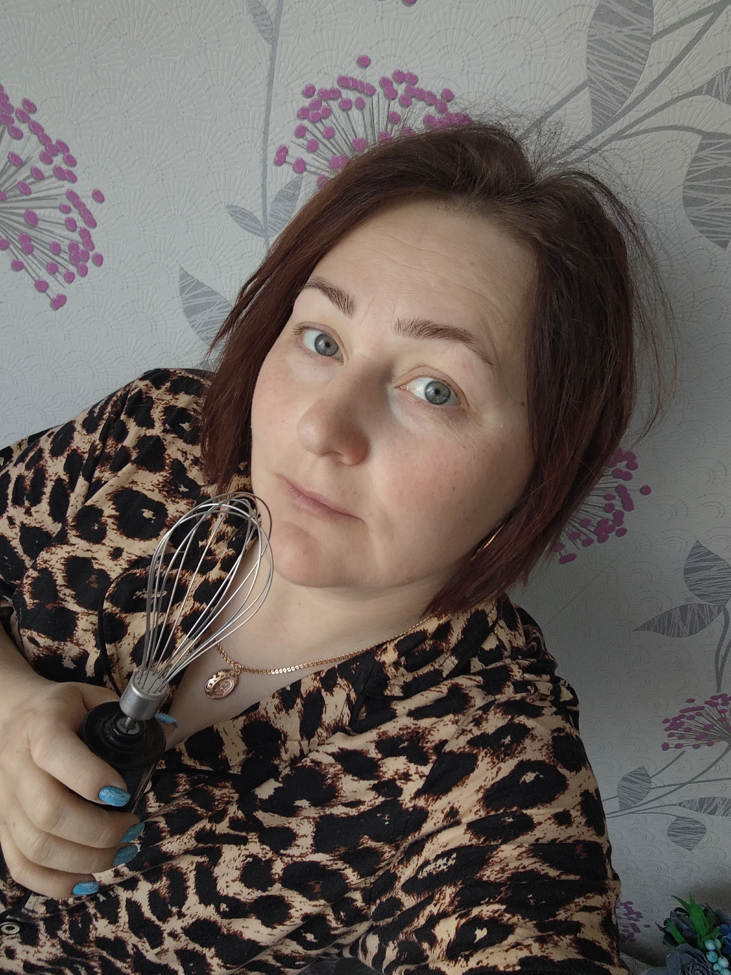 A medium shot shows a woman with auburn hair and blue eyes. She wears a tan and black leopard-print blouse and a gold necklace. She is holding a wire whisk with a black handle. The background is a patterned wallpaper with pink floral designs. The image was taken in Markovskii, Russia.