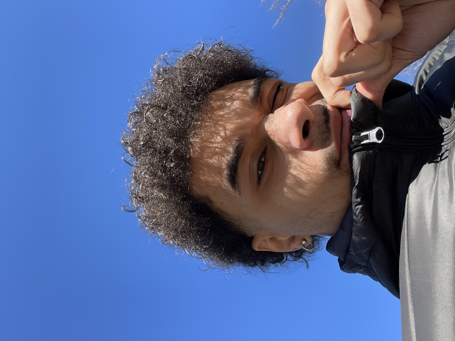 A close-up, low-angle portrait captures a male individual outdoors in Montigny-le-Bretonneux, France, against a clear blue sky. The subject, viewed from the chest up, has dark, curly hair with faded sides and a light mustache and goatee. He wears a black puffer jacket with a visible silver zipper pull. His right hand (viewer's left) is partially visible, pinching the jacket collar near his chin. His left eye (viewer's right) is squinted, while his right eye (viewer's left) is open, looking directly at the viewer. Dappled shadows, consistent with sunlight filtering through an overhead irregular structure or foliage, are cast across his face. A silver-colored hoop earring is visible in his left ear (viewer's right).