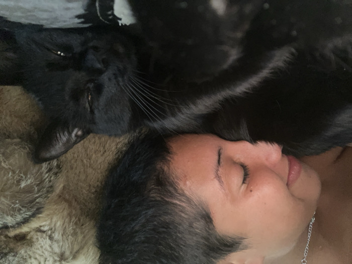 An indoor, close-up photograph displays one human individual and one black domestic cat. The human, a child or young person with light brown skin, short dark hair, and a silver chain necklace, occupies the left side of the frame. Their eyes are closed, and their mouth is slightly open, revealing the tip of the tongue, consistent with a state of sleep or deep rest. Their head is resting directly on the body of the black cat, which is visible on the right side of the frame. The cat's head is partially visible with eyes partially closed, and whiskers extended, also indicating a state of rest. The background consists of a textured, multi-toned brown and grey furry material. Below the cat's head, a dark patterned fabric is visible, consistent with bedding. The precise location within the city of Buenos Aires, Argentina, cannot be determined from the visual evidence.