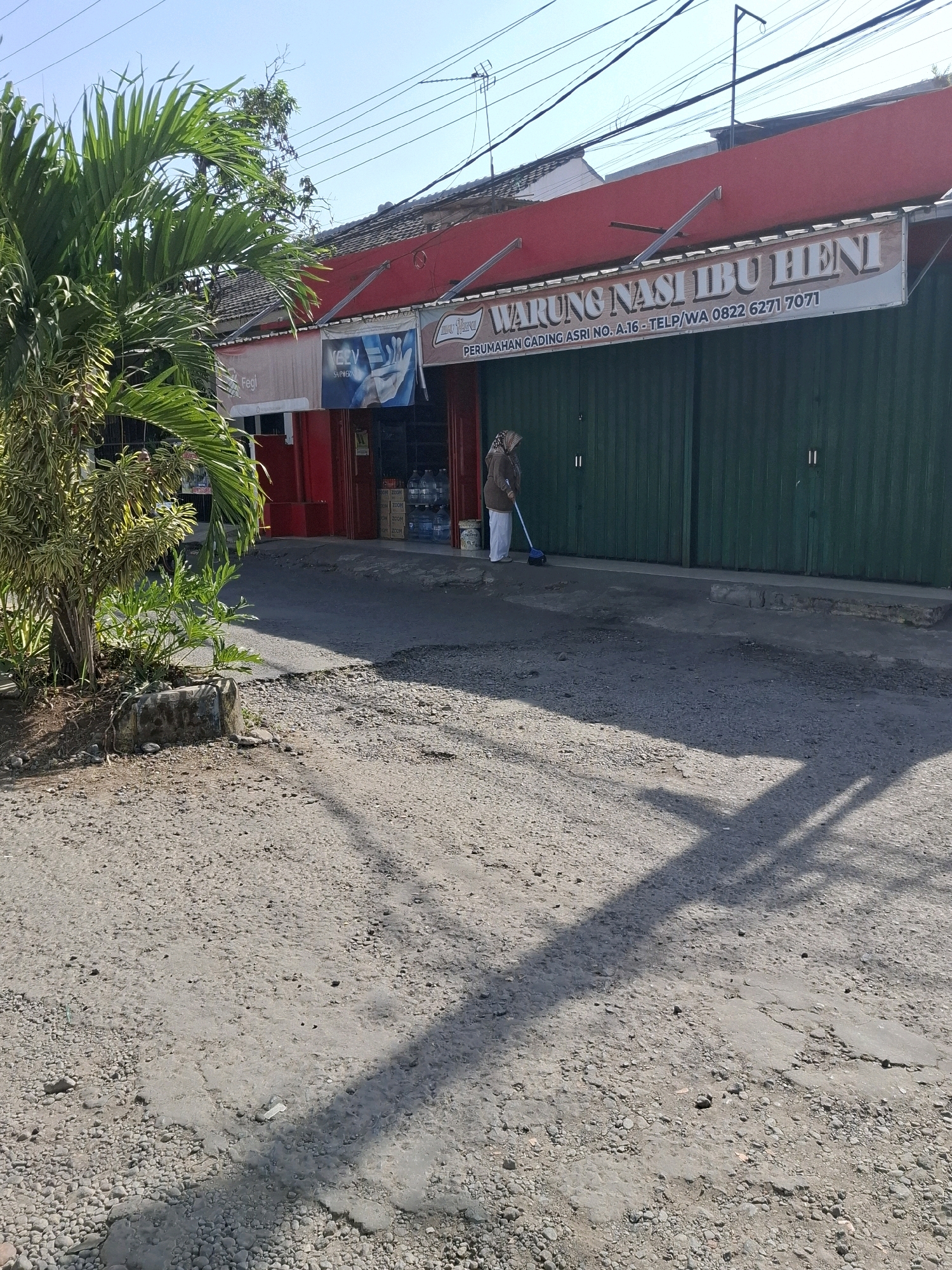 This is a medium shot, eye-level, outdoor, daytime, vertical photograph taken on a sunny day in the city of Karangtengah, Indonesia. In the foreground, a rough, gray asphalt road is visible, with small stones and patches of concrete. The road is partially covered by the shadow of a palm tree. To the left, a large palm tree and other green foliage are prominent.
In the midground, a woman is sweeping the ground with a blue broom. She is wearing a brown hijab and a long-sleeved brown top with white pants. Behind her, a building with a red facade and a sign that reads "WARUNG NASI IBU HENI" is situated. The sign also includes text about "PERUMAHAN GADING ASRI NO. A.16" and a phone number. The building has green corrugated metal doors and a tiled roof. In front of the building, several large bottles of water are stacked.
The background shows a clear blue sky with power lines and some tree branches. The overall scene depicts a street in a residential or commercial area, with everyday activity taking place.