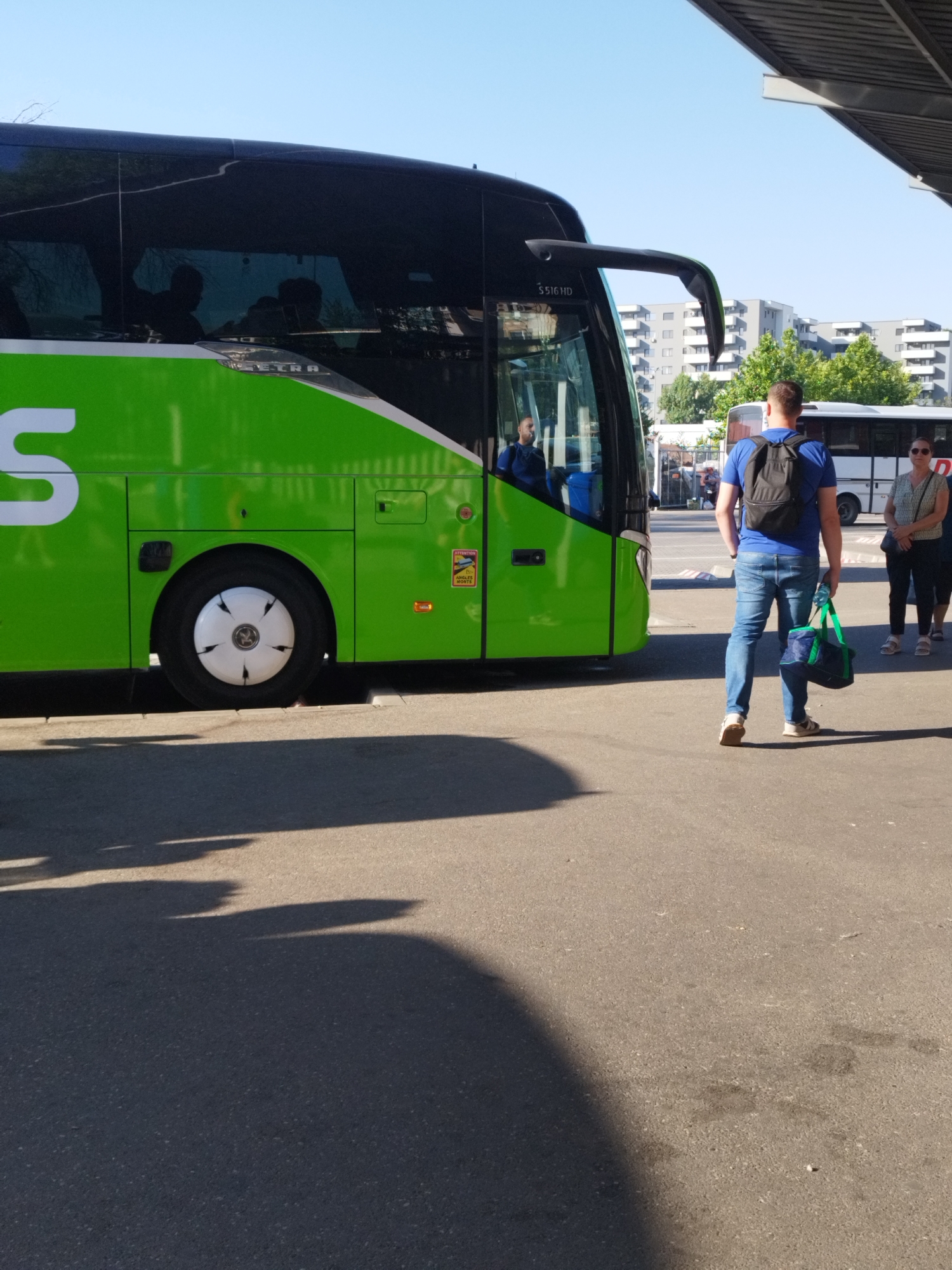 A green and black FlixBus coach is parked at a bus station. A man wearing a blue t-shirt, jeans, and a black backpack is walking away from the bus, carrying a green and black duffel bag. A woman in a patterned shirt and dark pants is standing to the right of the man. The bus station appears to be an open-air facility with a concrete ground surface. In the background, a white bus and several residential buildings are visible. The sky is clear and blue. The scene is brightly lit by sunlight, casting shadows on the ground.