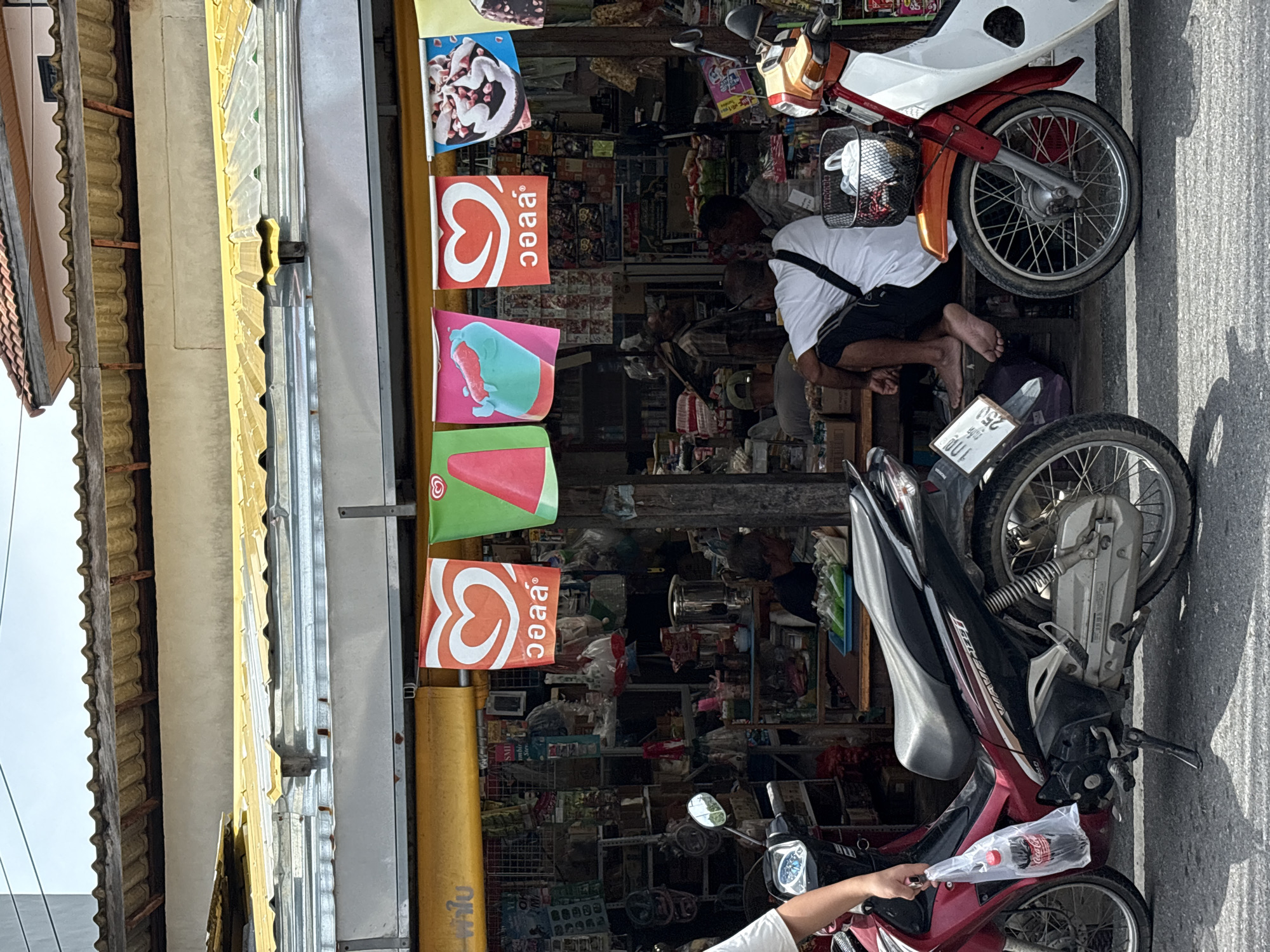 The image shows a street scene in Choeng Thale, Thailand, with a row of storefronts and parked motorcycles. In the foreground, a person on the left is holding a plastic bag containing a Coca-Cola bottle. To the right, a red and white motorcycle is parked. Behind the motorcycles, a storefront is visible, with a yellow awning extending over the entrance. Several people are seated or standing inside the store, which appears to be a small convenience store or market selling various goods. The storefront is decorated with colorful banners advertising ice cream. The overall impression is of a bustling street in a tropical climate.