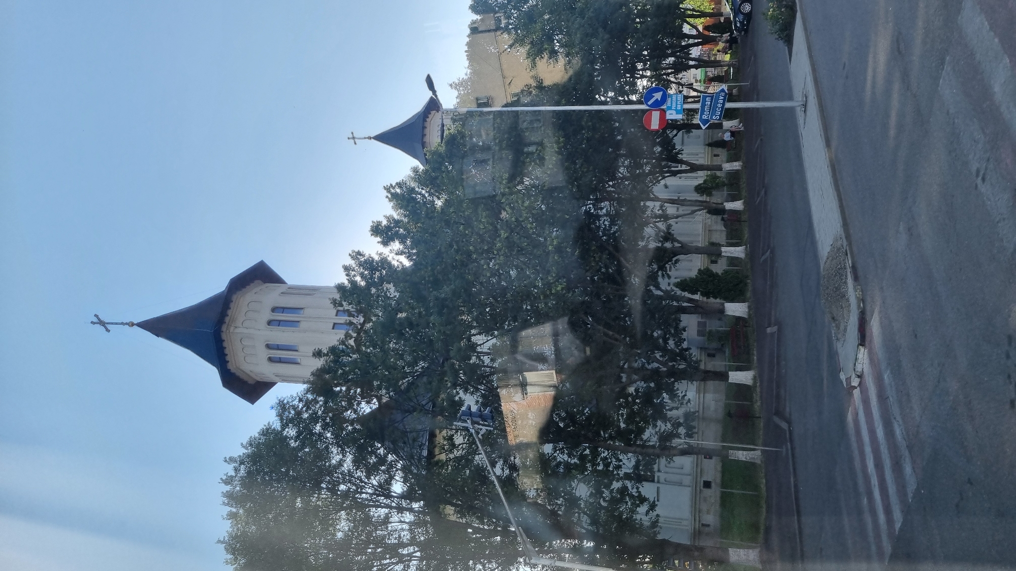 The image shows a view of the city of Fălticeni, Romania, focusing on a large building with two prominent towers, likely a church or monastery. The building has a white facade and dark, pointed roofs topped with crosses. Trees with white-painted trunks partially obscure the building. In the foreground, there is a street with a pedestrian crossing. Traffic signs are visible, including one pointing left with "Roman" and "Suceava" written on it, indicating directions to these cities. A blue sign above it reads "ZONA CU PLATA" which translates to "paid zone". A dark-colored car is parked on the right side of the street. The sky is clear and blue, suggesting it is daytime. There are no people clearly visible or engaged in any significant action. The scene is static and captures a moment of urban landscape.