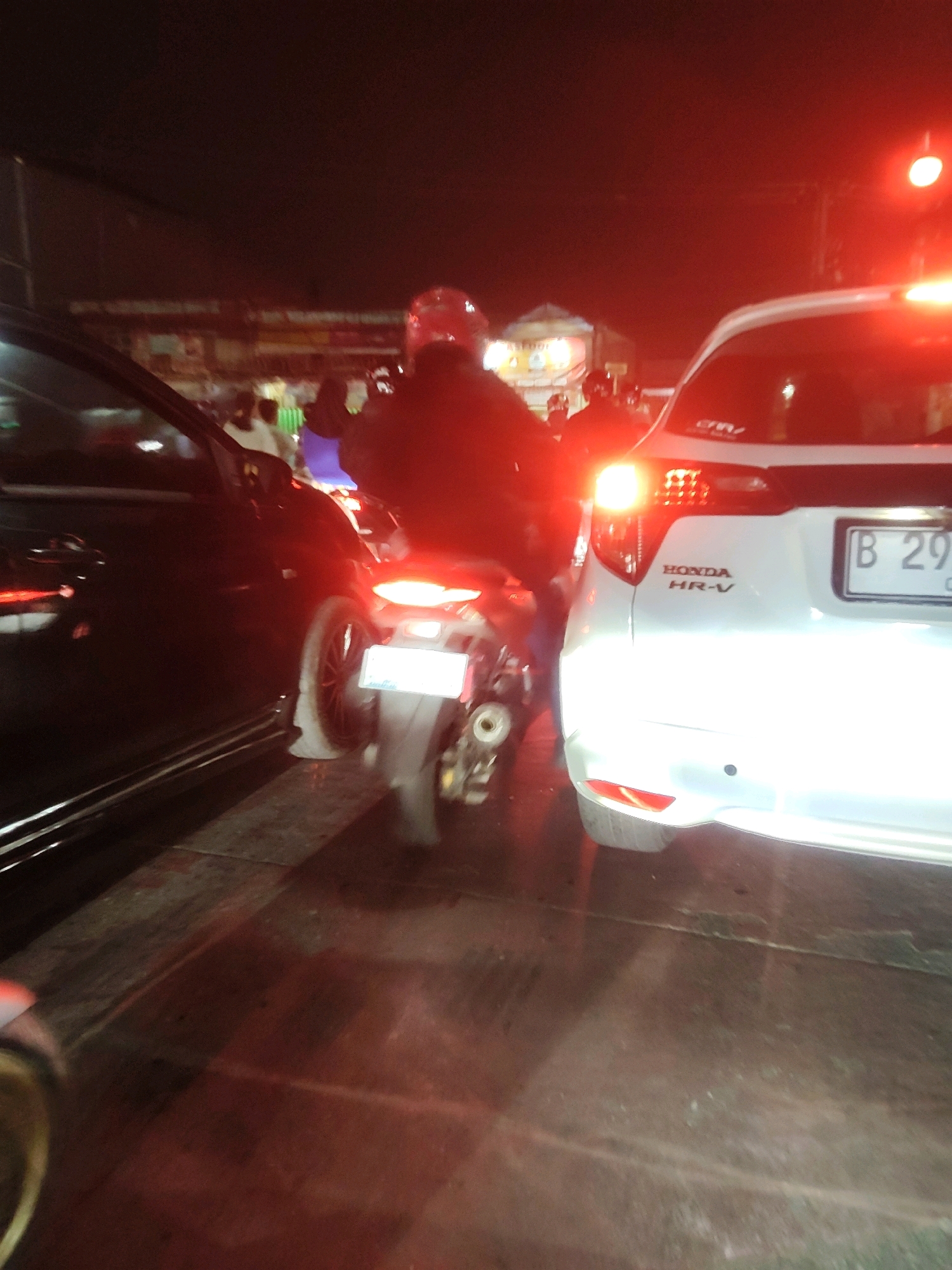 The image depicts a nighttime street scene in Mekarmukti, Indonesia, with several vehicles and pedestrians. A white Honda HR-V is parked on the right, displaying its rear license plate "B 29". To the left of the Honda, a black car is visible, partially obscuring a motorcycle. A person wearing a helmet is seated on the motorcycle, facing away from the camera. In the background, there are blurry figures of people and buildings, illuminated by streetlights and vehicle lights, suggesting a commercial or busy area. The overall lighting is low, with prominent red and white lights from vehicles and possibly traffic signals, creating a somewhat hazy atmosphere.