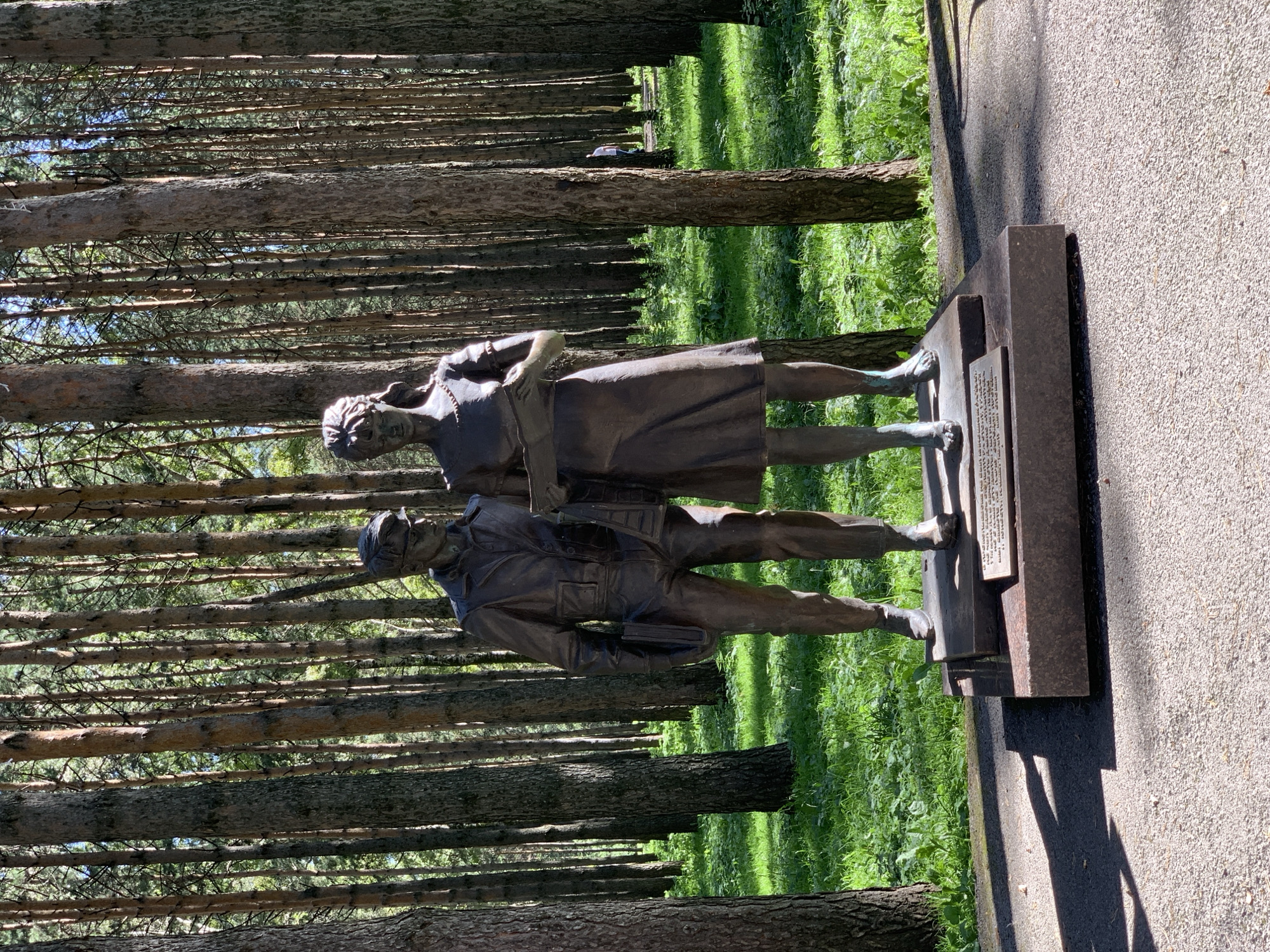 This image depicts a bronze sculpture of a man and a woman standing side-by-side in a pine forest. The man, on the left, wears a suit and glasses, and holds a book in his left hand. The woman, on the right, wears a dress and holds a book. They stand on a multi-tiered stone pedestal. Behind them, a dense forest of tall pine trees creates a backdrop, with sunlight filtering through the branches and illuminating patches of green grass. In the distance, a person walks among the trees. The sculpture is located in the city of Prokopyevsk, Russia, specifically at the monument to the poet and miner, Ivan Lysov.
