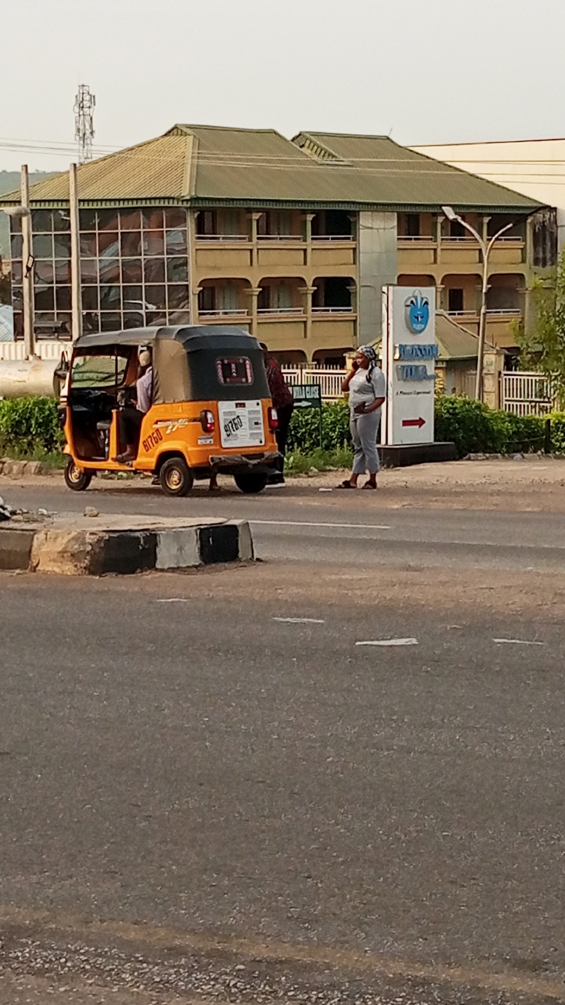 The image shows a street scene in Adankolo, Nigeria. An orange and black "BITGO" auto rickshaw (tuk-tuk) is stopped on the side of the road near a curb. A woman wearing light-colored clothing and a head covering is standing near the auto rickshaw, appearing to be on her phone. There is another individual standing nearby the tuk-tuk.
In the background, a multi-story building with a green roof can be seen. A sign that reads "REMSON HOTELS" can also be seen in the background to the right of the building. The sky is overcast. The road is asphalt and shows lane markings.