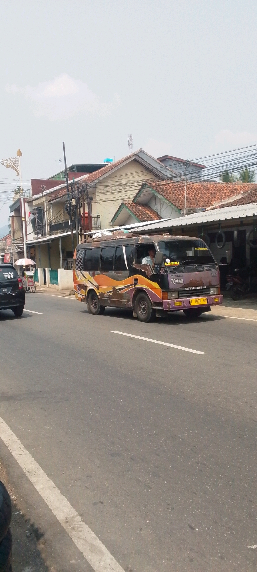 The image shows a street scene in Sukasari, Indonesia. A slightly dilapidated, purple and orange decorated minibus, identified by the name "Raisa" and the license plate "F 7893 HA", is driving on the right side of the road. The minibus has a driver visible inside. To the left of the minibus, a black car is also traveling in the same direction. Further to the left, a vendor with a cart, possibly selling food or drinks, is visible on the side of the road. The background features buildings with tiled roofs, power lines, and a cloudy sky. The road surface is asphalt with white lane markings.
