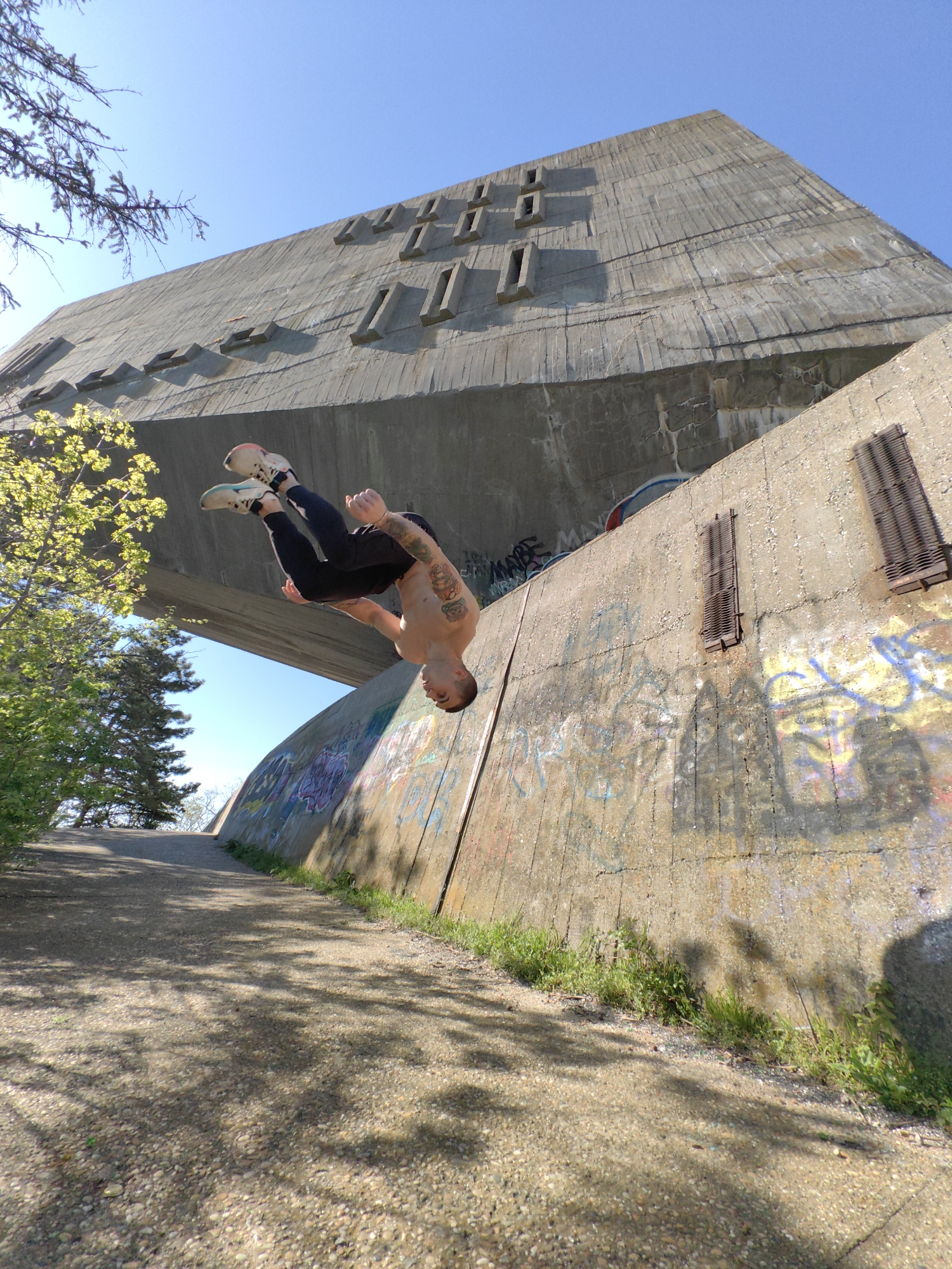 A shirtless man in dark pants and sneakers is performing a backflip near a large concrete structure in Varna, Bulgaria. The structure is characterized by a rectangular shape with evenly spaced, protruding rectangular blocks along its upper section. The structure features heavily graffitied concrete walls with metal ventilation grids. A pathway covered in gravel and dirt runs alongside the structure. Sunlight streams through branches, creating shadows on the ground. Some green vegetation is visible. The sky is clear and blue.