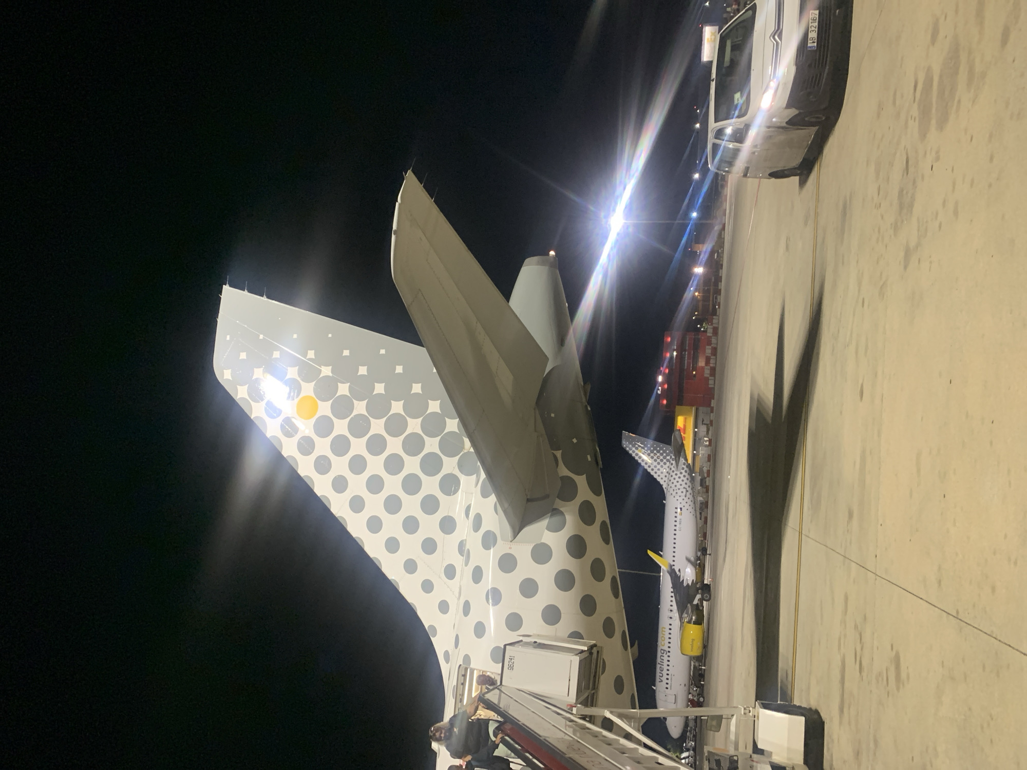 At night on an airport tarmac in Sant Boi de Llobregat, Spain, a Vueling Airlines airplane is parked. Its tail, decorated with a pattern of gray polka dots and a yellow circle, is prominently featured in the foreground. A set of mobile stairs is attached to the side of the aircraft, with several people ascending and descending. In the background, another Vueling airplane and a yellow and red structure are visible. A white van, with the license plate WB 32167, is parked on the tarmac to the right of the frame, illuminated by the aircraft's lights. The scene is lit by various airport lights, creating reflections and lens flares.