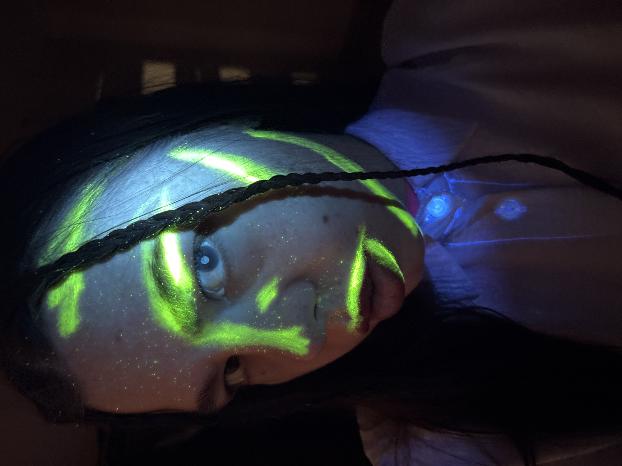The image shows a close-up of a person's face, tilted at an angle. The person has fair skin and dark hair, with one braid crossing the forehead. A green light pattern is projected onto their face. The light appears to be a stylized "K". There are small points of green light on the skin as well. The person's left eye is visible and appears to be light blue. A glimpse of a collar with two visible light fixtures is at the person’s neck. The background is dark. The location of Perm, Russia is not identifiable from the image.