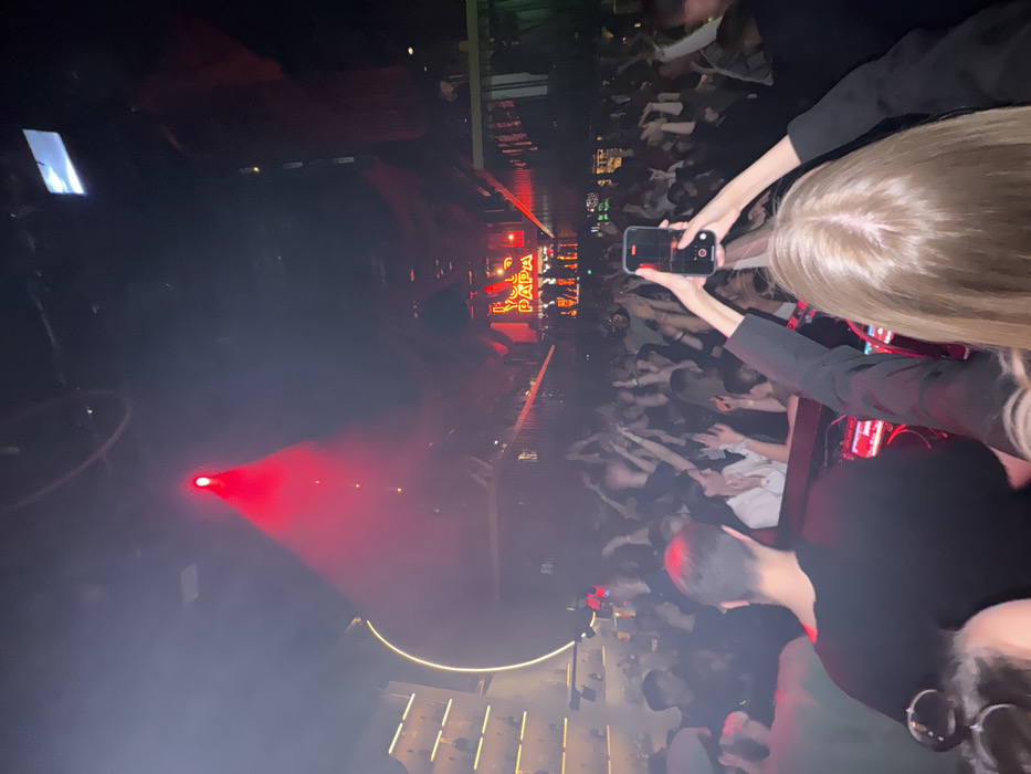 The image depicts the interior of the MAMA RUSSIA PAPA bar, located at Nikoloyamskaya St, 28/8, Moscow, Russia, during a crowded event. A large audience fills the central area, with many individuals raising their arms, indicating active engagement.
In the foreground, the back of a person with light-colored hair, wearing a dark long-sleeved top, holds a black smartphone vertically, actively recording the scene. The back of another individual's head, with short dark hair and a black t-shirt, is also partially visible.
In the mid-ground, a DJ booth is visible with electronic equipment, and a DJ is present, facing the crowd. Behind the DJ, a stage area features a prominent red neon sign displaying the establishment's name, "MAMA RUSSIA PAPA." Additional figures, likely performers or dancers, are discernible on the stage.
The environment is characterized by dark overhead structures and intense red stage lighting emanating from spotlights, creating visible atmospheric haze. On the left side of the frame, a bar area with illuminated shelves stocked with bottles is present, along with a large, vertically curved illuminated panel. Elevated sections with railings are visible on the right, indicative of a multi-level entertainment venue.