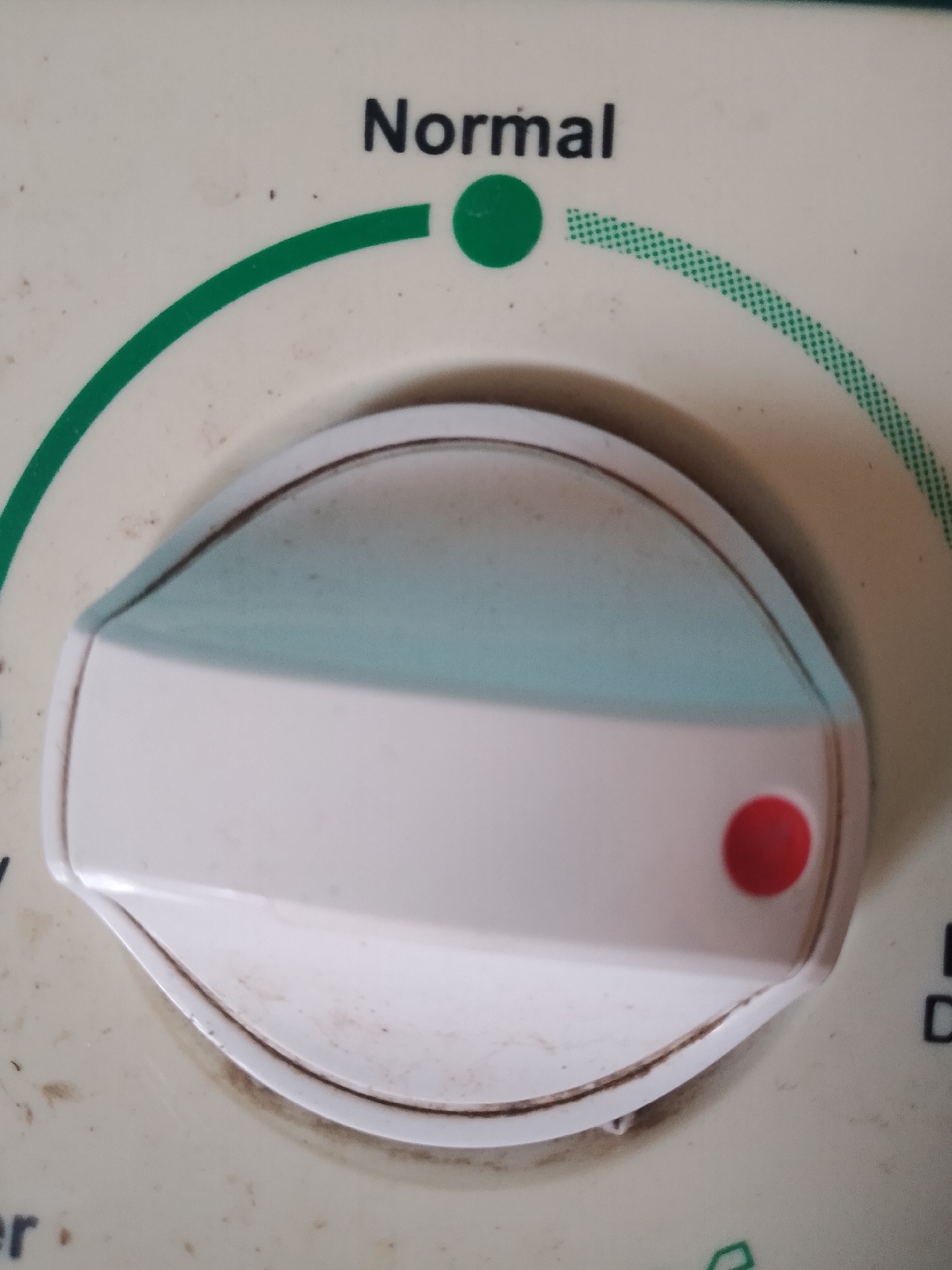 The image shows a close-up of a control knob on a cream-colored appliance. The knob is white with a light blue gradient on its top surface and a red dot on its front. Above the knob, the word "Normal" is printed in black text. A green arc with a dashed green line runs around the knob. The appliance appears to be dirty with some brown specks and smudges. The background is out of focus. There are no people or specific environmental details visible to determine a location in Santa Ana, Venezuela.