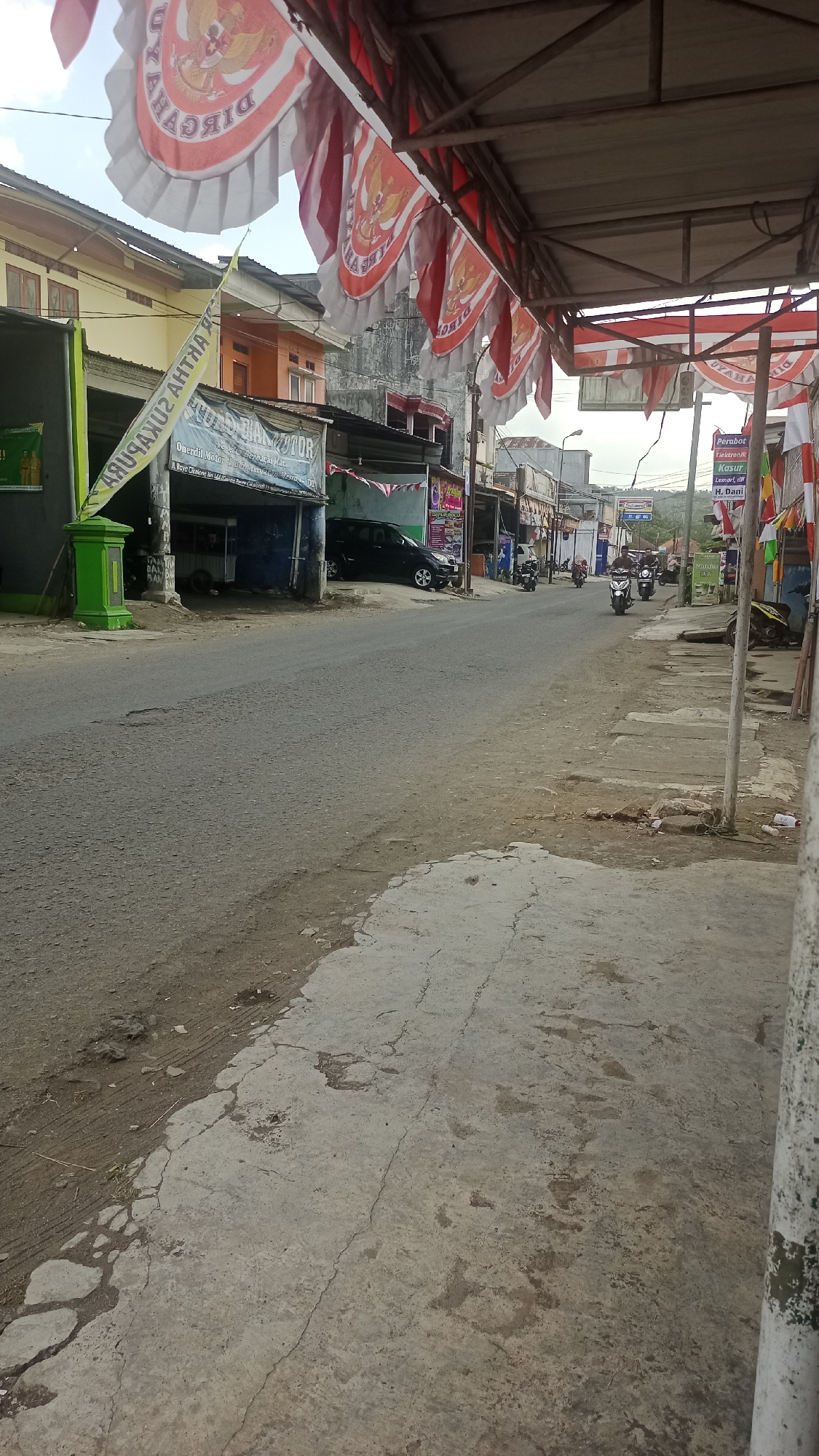 The image depicts a street scene in Cikalong, Indonesia, likely during the day with overcast conditions. The street is paved with asphalt and shows signs of wear and tear, with cracks and patches visible. The foreground features a cracked concrete sidewalk with dirt and debris scattered on it.
On the left side of the street, several buildings are visible. One building has a green pillar in front of it and a banner that appears to be advertising a business. Further down, there's a dark-colored vehicle parked. A banner with red and white motifs and images of eagles is strung across the top of the scene, suggesting a national celebration or event.
On the right side of the street, more buildings with shop fronts line the road. One shop has a sign that reads "Perabot Elektronik, Kasur, Lemari, dll" which translates to "Electronic Furniture, Mattress, Wardrobe, etc." Another sign is partially visible with the name "H. Dani." Several motorcycles are present, with some parked and others being ridden on the street. The background shows more buildings and a hilly landscape under a cloudy sky.
There are several people visible in the image, mostly riding motorcycles. Their presence indicates ongoing daily activities. The overall impression is of a busy, functional street in a local Indonesian town.