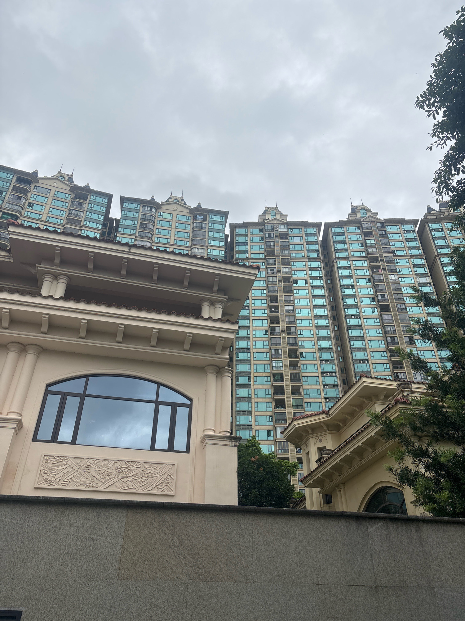 In Nanhai District, China, a low-angle perspective captures a cluster of modern residential high-rises with blue-tinted windows against a cloudy sky. In the foreground, a beige, classically inspired building with arched windows and ornate detailing sits behind a grey stone wall. Lush green trees are interspersed throughout the scene, framing the buildings. The environment appears to be a well-maintained urban residential area, with no people or vehicles visible. The image is a static, detailed snapshot of an architectural landscape.