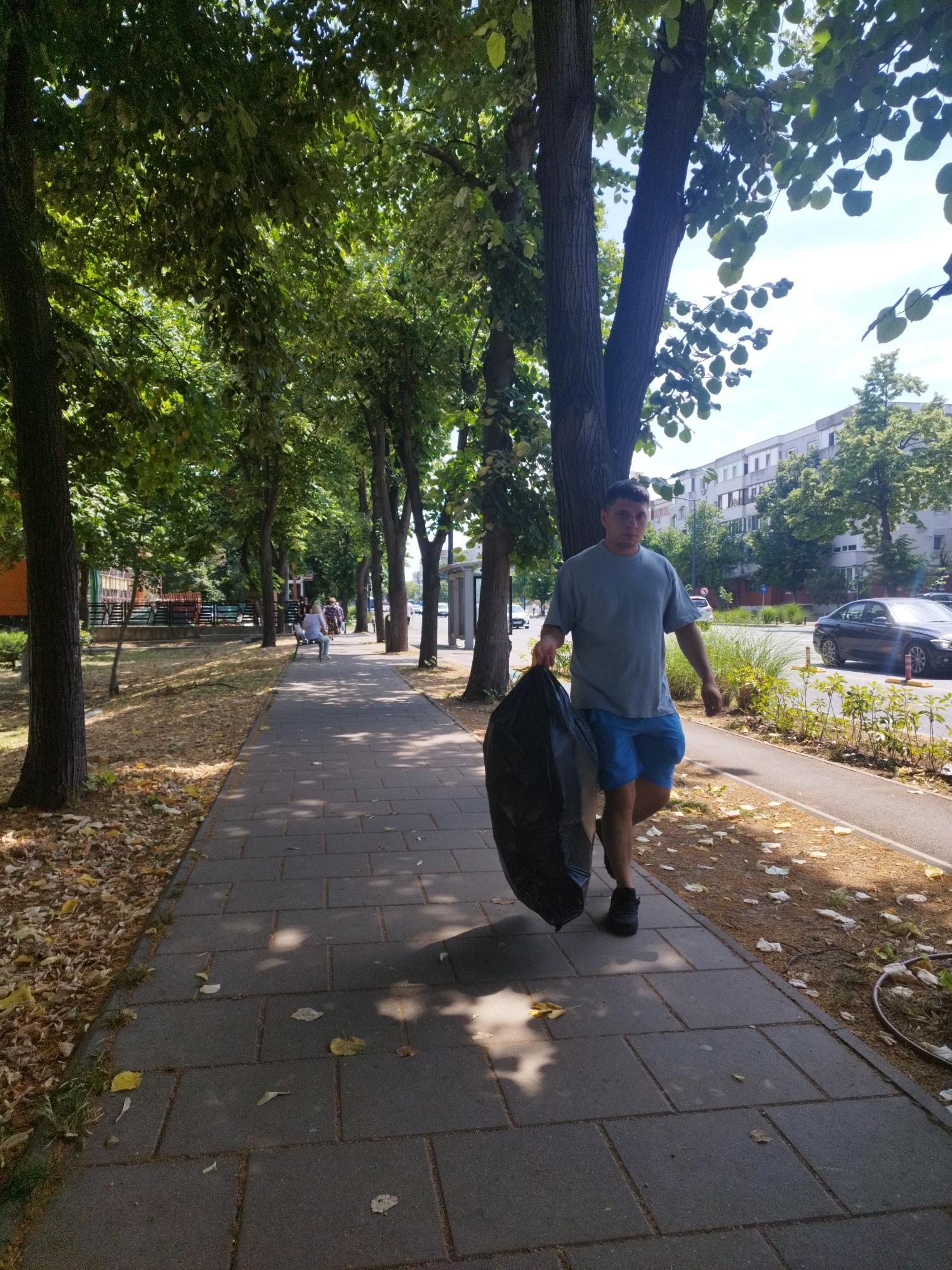 A man is walking on a paved sidewalk carrying a large black trash bag. He is wearing a grey t-shirt and blue shorts, with black shoes. The sidewalk is lined with trees on the left and a street with a black car and a white building in the background on the right. The ground on the left of the sidewalk is covered with dry leaves. Several other pedestrians are visible further down the sidewalk. The scene appears to be in a city, likely Constanța, Romania given the architecture and street layout.