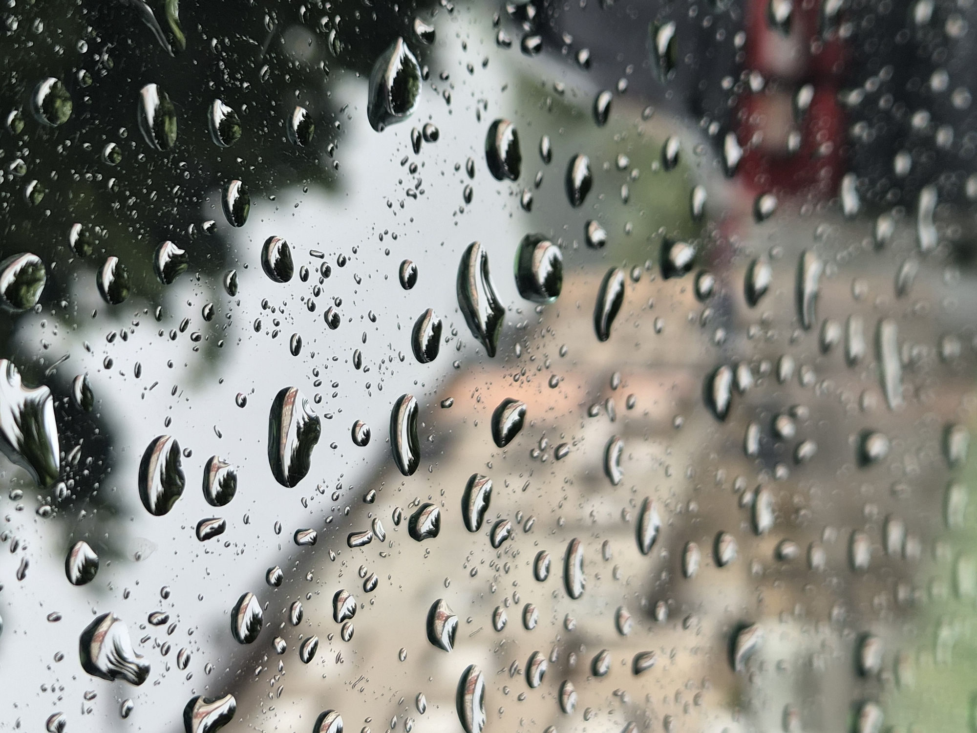 The image presents a close-up view of a glass surface, likely a window, adorned with numerous water droplets of varying sizes. The drops are sharply focused, creating a shallow depth of field that renders the background in a soft, abstract blur (bokeh effect). This indicates a rainy or post-rainy condition in Differdange, Luxembourg.
The water droplets act as tiny lenses, reflecting and refracting the blurred scene beyond. Within many of the larger drops, distorted and inverted impressions of the background are visible, including hints of dark green foliage, lighter grey/beige structures, and a distinct, blurred red object towards the mid-right. The overall background suggests an urban or suburban environment with trees and buildings.
The lighting appears to be diffused, consistent with an overcast daytime setting during precipitation. The image focuses on the tactile and visual texture of the wet glass. There is no visible text present in the media file.