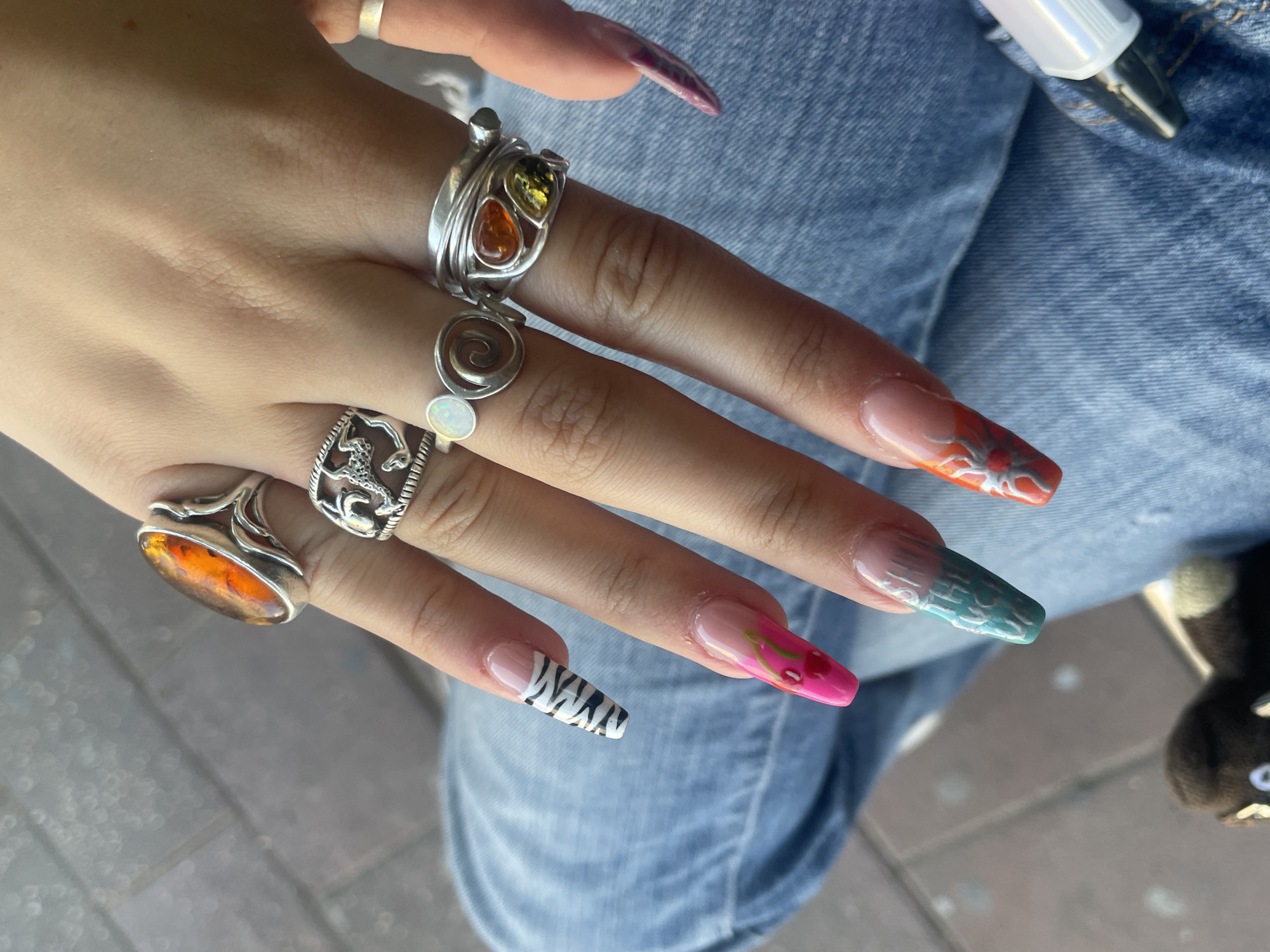 A close-up, overhead shot features a person's hands adorned with multiple silver rings and elaborate, long, artificial fingernails. The rings are varied in design, with one prominent large oval amber stone, another featuring a detailed sea creature, a small white opal, and a cluster of teardrop-shaped amber and green stones. The fingernails display a range of artistic designs: one has a black and white zebra stripe pattern, another is a bright pink with cherries, a third is a teal gradient with white text, and the furthest right is orange with a silver floral design. The background suggests an outdoor setting with a textured grey surface resembling pavement and the blue denim of jeans. The scene is likely situated in Ramat Gan, Israel, given the provided location context.