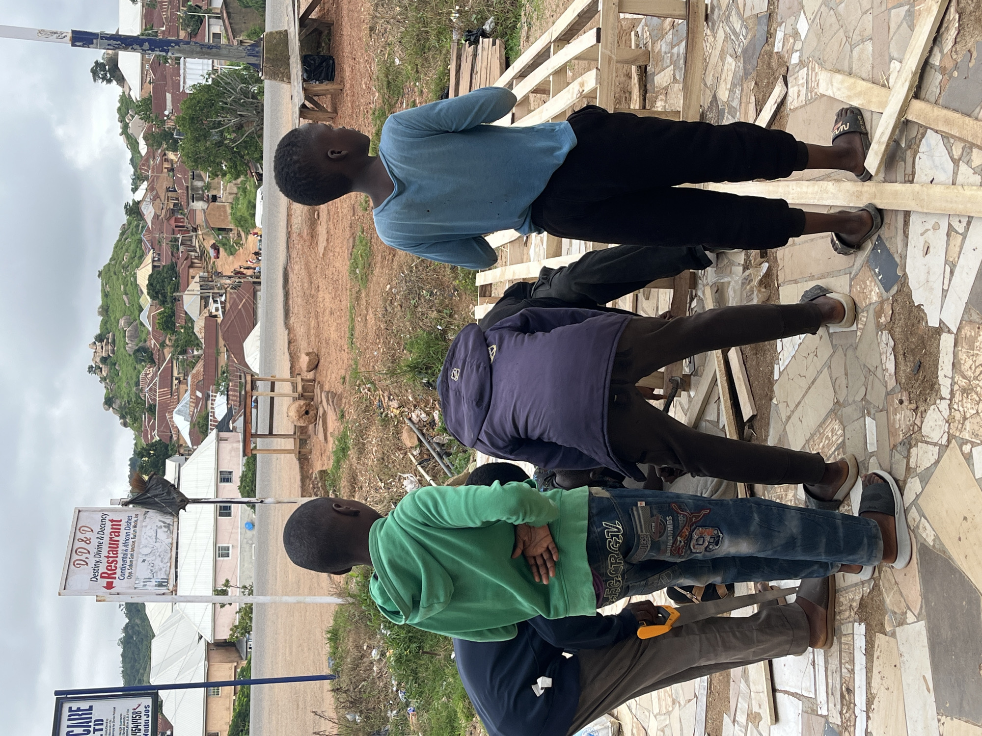 In Jos, Nigeria, on a cloudy day, several young boys are gathered around a construction project on a paved area. One boy in a green hoodie stands with his hands behind his back, observing. Another boy in a blue long-sleeved shirt is positioned to the right, also looking away. Two other boys are bent over, seemingly engaged in the construction. One of them is holding a saw, and the other is wearing a dark purple hooded jacket. Scattered wooden planks are visible on the ground, indicating they are working with wood. In the background, a village with red-roofed houses is nestled on a hillside, with rocky outcrops and green vegetation. A sign for "D.D & D Restaurant" is prominently displayed, along with other signs for "CARE LTD" and other businesses, suggesting the location is near a commercial area. The overall environment appears to be a mix of urban and semi-rural, with construction materials, debris, and natural elements.