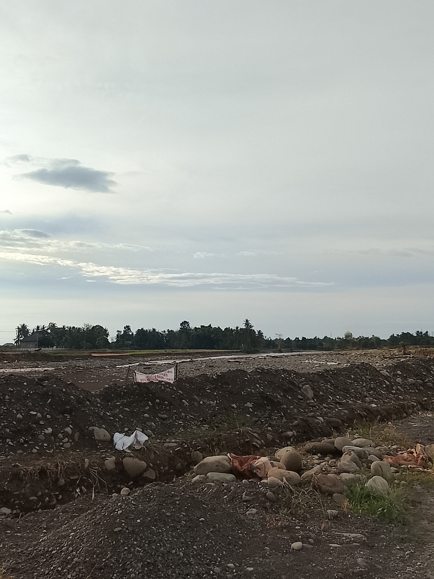 The image depicts a desolate, rocky landscape under an overcast sky, likely in Geunteng, Indonesia, given the context. The foreground is dominated by mounds of dark soil and scattered rocks, suggesting a construction or excavation site. A white plastic bag lies discarded amongst the rocks in the middle ground, and further back, a white banner with red lettering is partially visible, obscured by dirt and debris. Beyond this, a line of lush green trees marks the horizon, interspersed with a few buildings, including what appears to be a house with a red roof on the left and a domed structure further to the right. A river or body of water is visible between the foreground and the tree line, reflecting the muted sky. The overall impression is one of unfinished work and a somewhat neglected environment.