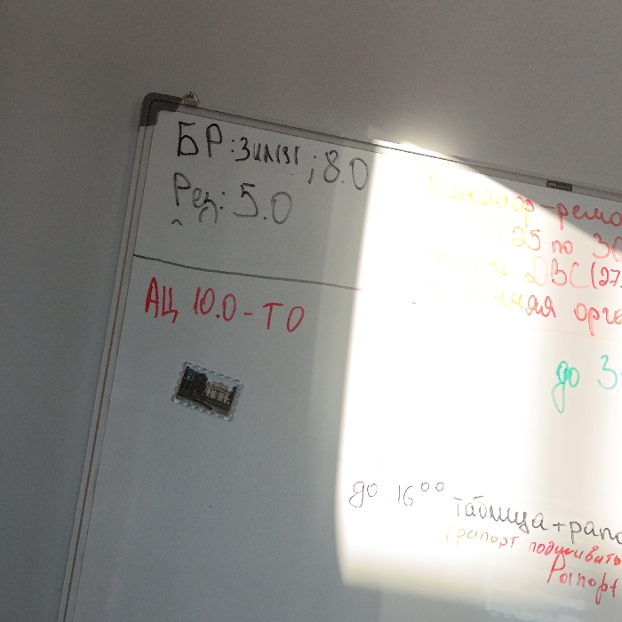 The image displays a white dry-erase board mounted in an indoor setting in Hlybokaye, Belarus. The board is positioned at an angle, tilting upward from left to right. A strong, diagonal glare from a bright light source significantly illuminates and obscures the right half of the whiteboard.
On the upper left portion of the board, two lines of text are written in black marker: "БР: ЗАКЛ; 8.0" and "Ред: 5.0". Below this, in red marker, is the text "АЦ 10.0-ТО". Directly beneath the red text, slightly left of center, a small rectangular object resembling a postage stamp is affixed. This object depicts a historical building, possibly a church or a palace, characterized by a prominent tower or spire.
The right side of the whiteboard is largely washed out by the glare, making much of the text illegible. However, fragmented words and numbers in various colors—yellow, orange, green, and red—are discernible. These include segments such as "...ремон...", "25 по 30", "...BC(27)", "...ная орг...", and "до 3..." in the upper and middle right sections. Towards the bottom right, below the glare, additional text written in black and red markers is visible: "до 16°°", followed by "Таблица + рано...", and a parenthetical phrase "(рапорт подписать)", with "Ронпорт" below it. No individuals are present in the scene.