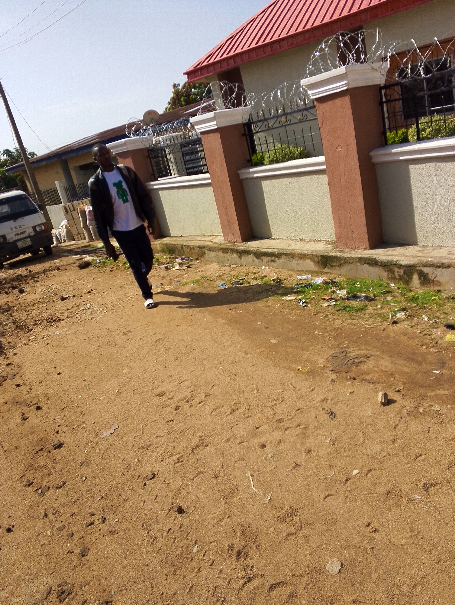 In Jos, Nigeria, a man in a black jacket and white t-shirt walks on a dirt road. The road is lined with a light-colored wall and a red-roofed building. The wall has a decorative fence topped with barbed wire. Debris and grass are scattered along the base of the wall. A white van is parked on the left side of the road. The sky is clear and blue.