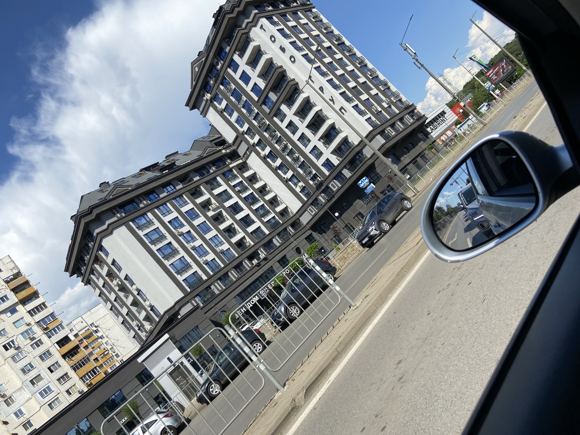 The image is a street-level, slightly tilted view of a modern residential building in Sofia, Bulgaria, on a sunny day with scattered clouds. The building, with its gray facade, dark trim, and numerous blue-tinted windows, dominates the right side of the frame. On the left side, an older, light-colored residential building with yellow accents is visible. In the foreground, a car's side mirror reflects the road and other vehicles. Cars are parked along the street in front of the buildings, separated from the road by a metal fence. A street sign is partially visible on the left. The overall impression is of an urban environment in the midst of development, with a mix of older and newer architecture.