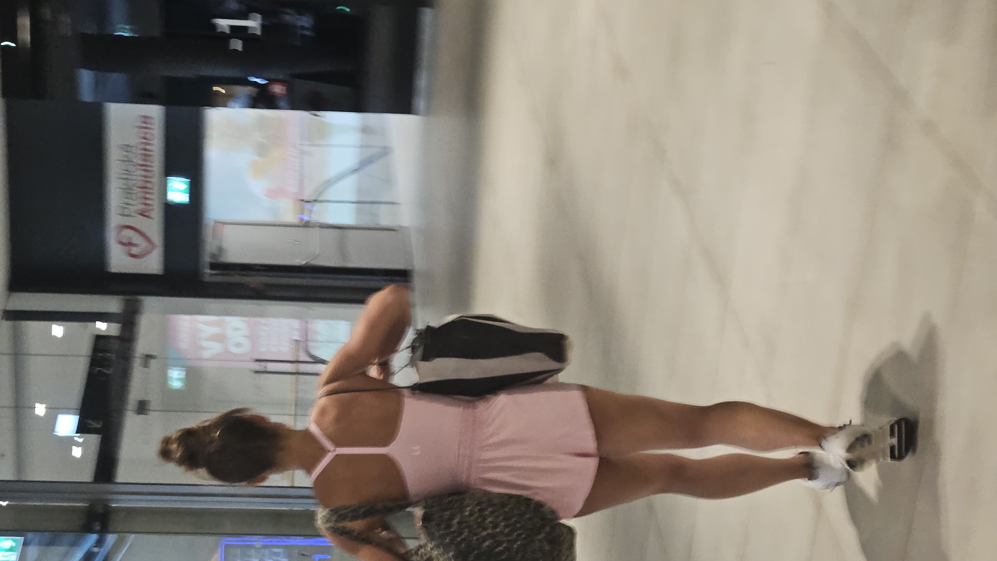 A woman with her back to the camera is walking away from the viewer in a brightly lit indoor space, possibly a mall or an airport terminal. She is wearing a pale pink athletic outfit, consisting of a tank top and shorts. She is carrying two large bags: one with a leopard print design in her left hand, and another with black and white stripes in her right hand. Her hair is pulled up in a bun. Her legs are visible from the mid-thigh down, and she is wearing white athletic shoes. In the background, there are signs and architectural elements of a modern building. One sign is illuminated and reads "Praktická Ambulancia", indicating a practical medical clinic. Another sign displays numbers "2-3". The floor is tiled in a light, neutral color. The overall impression is of someone on the move, possibly after a workout or during travel. The presence of the "Praktická Ambulancia" sign suggests the location could be in a facility that includes medical services, which might be found in a larger public complex in a city like Prešov, Slovakia.