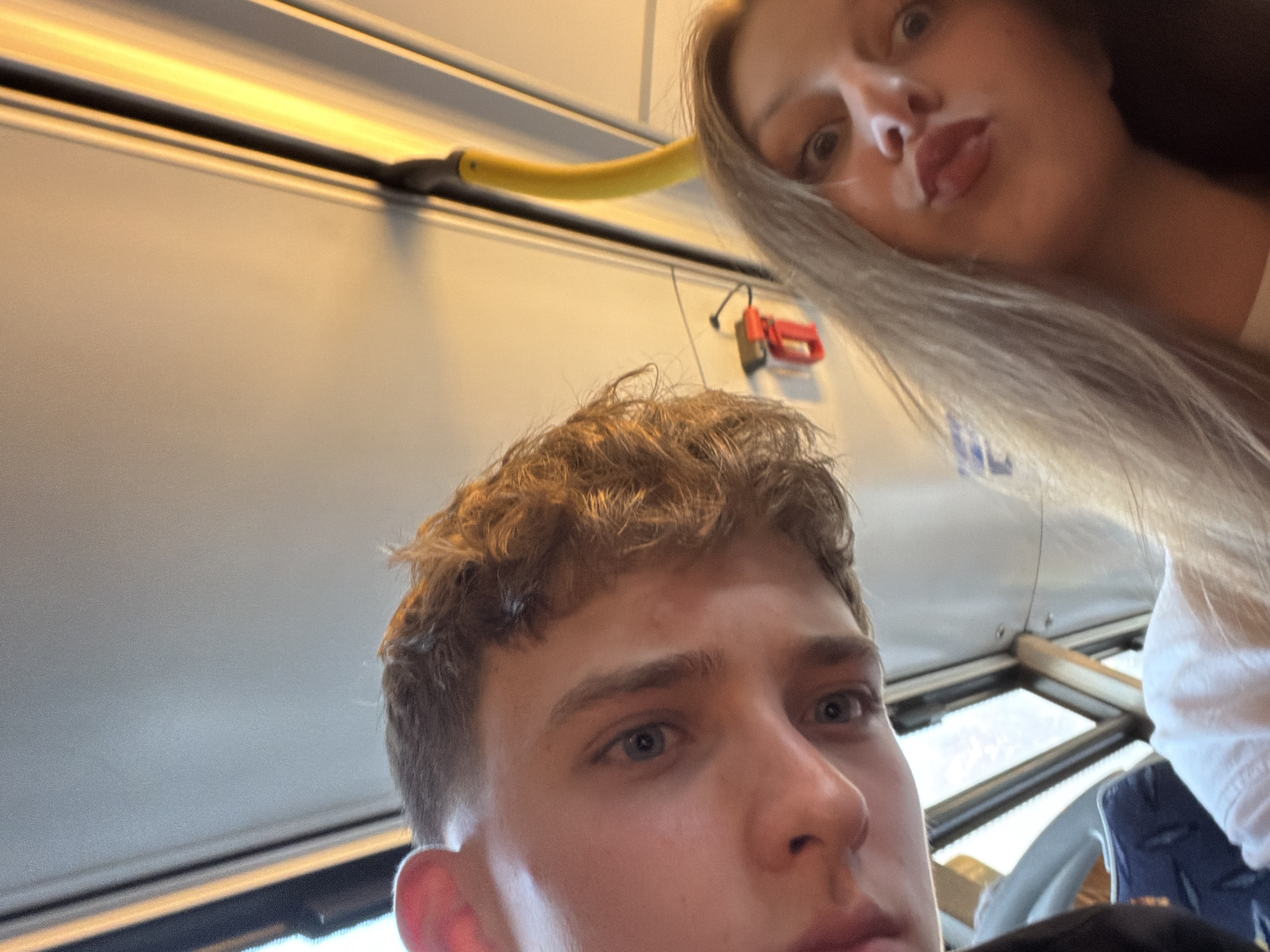 The image is a selfie taken from a low angle, showing the faces of two young people, likely a couple. The male on the left is closer to the camera, with light brown hair and blue eyes. The female on the right is slightly further away, with long blonde hair and is puckering her lips. They appear to be indoors, possibly on public transportation, indicated by the presence of a yellow pole and a window with a view of what appears to be urban infrastructure. The lighting suggests it is either morning or evening. The background is blurred, but elements of what could be seats and railings are visible. The overall scene is casual and personal, characteristic of a selfie taken during a journey. The location of Burgas, Bulgaria, cannot be determined from the visual information present in the image.