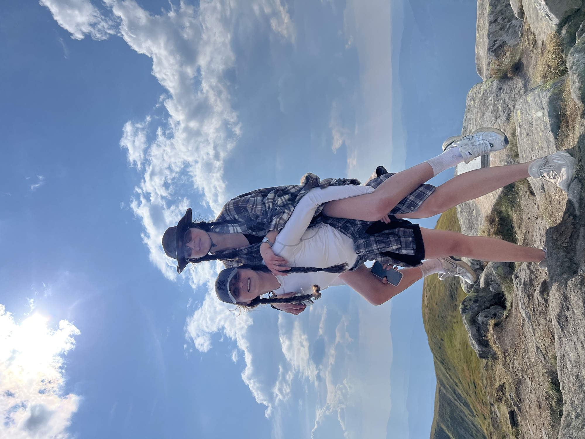 Two young women are on a mountain summit in Luhy, Ukraine, with a bright blue sky and scattered clouds above. The woman in front is giving the woman behind her a piggyback ride. Both women are wearing casual, outdoor clothing. The woman in front has on a white long-sleeved shirt, black shorts, white socks, and white sneakers. She is also wearing a grey baseball cap. The woman behind her is wearing a plaid shirt, sunglasses, and a hat. They are standing on a rocky surface with a scenic view of rolling hills and mountains in the distance. The sun is shining brightly, creating a slightly hazy effect in the sky.