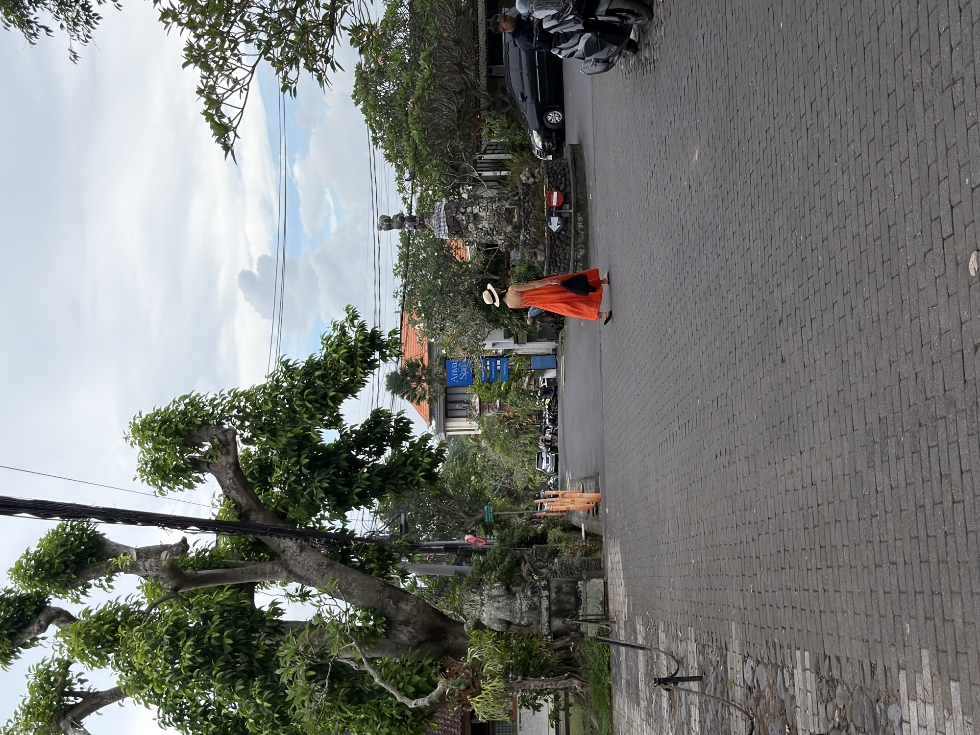 The image captures a daytime scene along a paved street or pathway in the Ubud District, Indonesia, under an overcast sky with patches of blue and white clouds. Lush green trees with dense foliage, some appearing gnarled and ancient, line both sides of the street, with power lines crisscrossing overhead.
In the mid-ground, a person is walking away from the viewer on the dark gray, block-paved surface. They are wearing a long, flowing orange or reddish skirt, a light-colored top, and a wide-brimmed straw hat. They carry a black handbag over their shoulder. Adjacent to their path, on the left, stands a traditional stone structure, possibly a shrine or monument, partially wrapped in a distinctive black and white checkered fabric (poleng cloth). Nearby, a red circular sign with a white bar (a "no entry" or similar traffic sign) is visible.
Further down the street, several motorcycles or scooters are parked or moving, and a dark car is also visible. Buildings with traditional Balinese architectural elements, including orange-tiled roofs, are visible in the background. On one building, blue signs are visible, one of which clearly reads "Anvaya Spa." Orange traffic cones are also visible on the side of the road in the distance. The overall atmosphere suggests a relaxed, tropical setting.