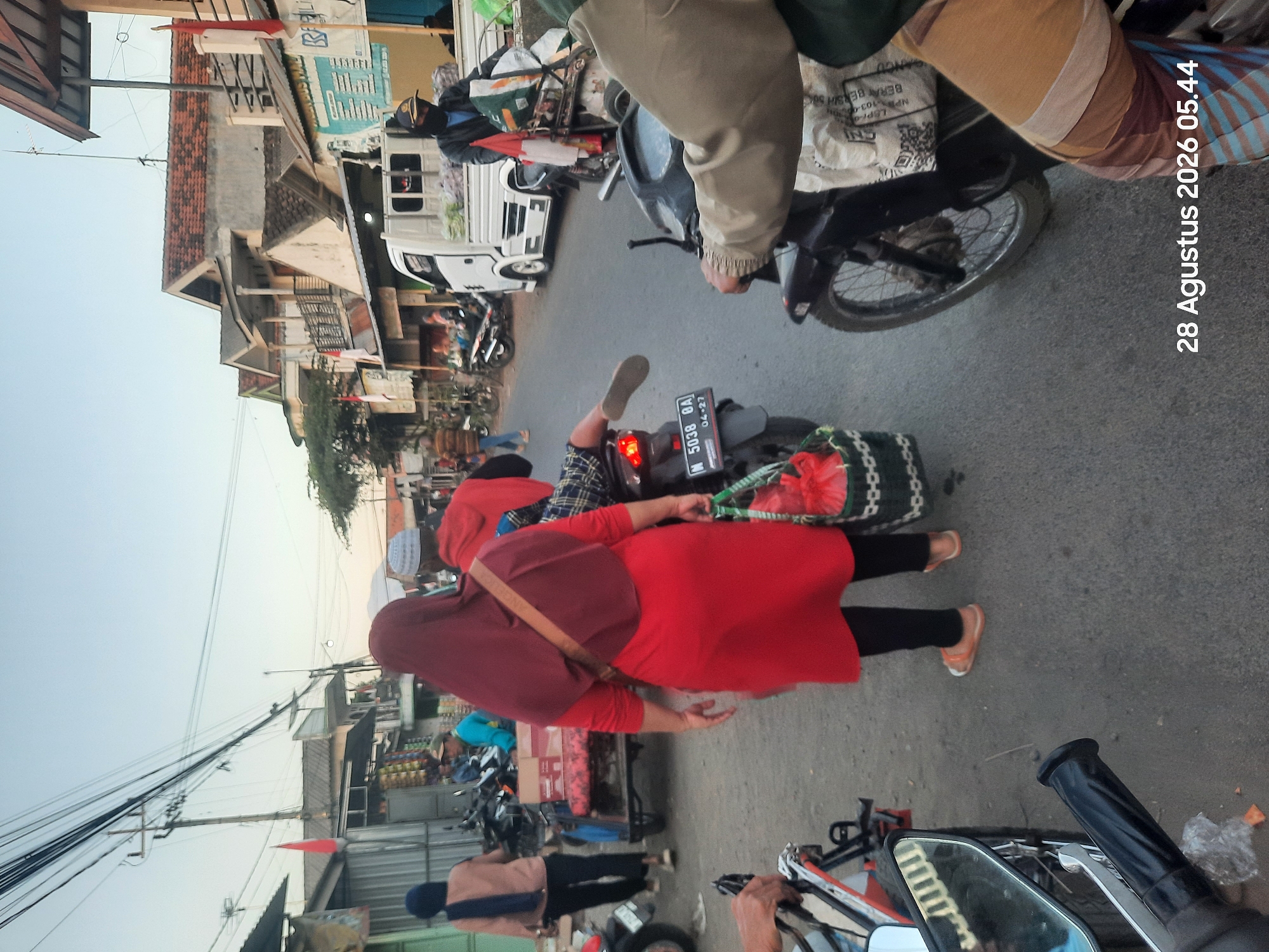 The image depicts a street scene in Pajarakan Kulon, Indonesia, on August 28, 2026, at 05:44. The sky is overcast, suggesting either early morning or late evening. A woman wearing a red hijab and a red dress, carrying a green woven bag with red items inside, walks away from the camera down a paved street. She wears orange sandals. To her left, a person in a light brown jacket walks away from the camera, wearing dark trousers and dark shoes. In the background, several buildings with reddish-brown tiled roofs line the street, some appearing to be shops or residences. Power lines are strung across the sky, supported by poles.
Several motorcycles are present. One is in the foreground, partially visible, with its handlebars and mirror showing. Another motorcycle is ridden by a person in a tan jacket and plaid shirt, who is seen from the waist down, with one foot resting on the motorcycle's rear fender, their leg extended backward. This motorcycle has a license plate reading "M 5038 GA" and "04-27". To the right of this motorcycle, another motorcycle is parked, carrying various goods, including what appears to be produce. Behind this, a white truck with a flatbed is parked, also carrying goods. Further down the street, more motorcycles and a cart are visible. A red flag on a pole is seen to the right of the white truck. The overall scene suggests a busy street, possibly a market area, with people and vehicles engaged in daily activities.