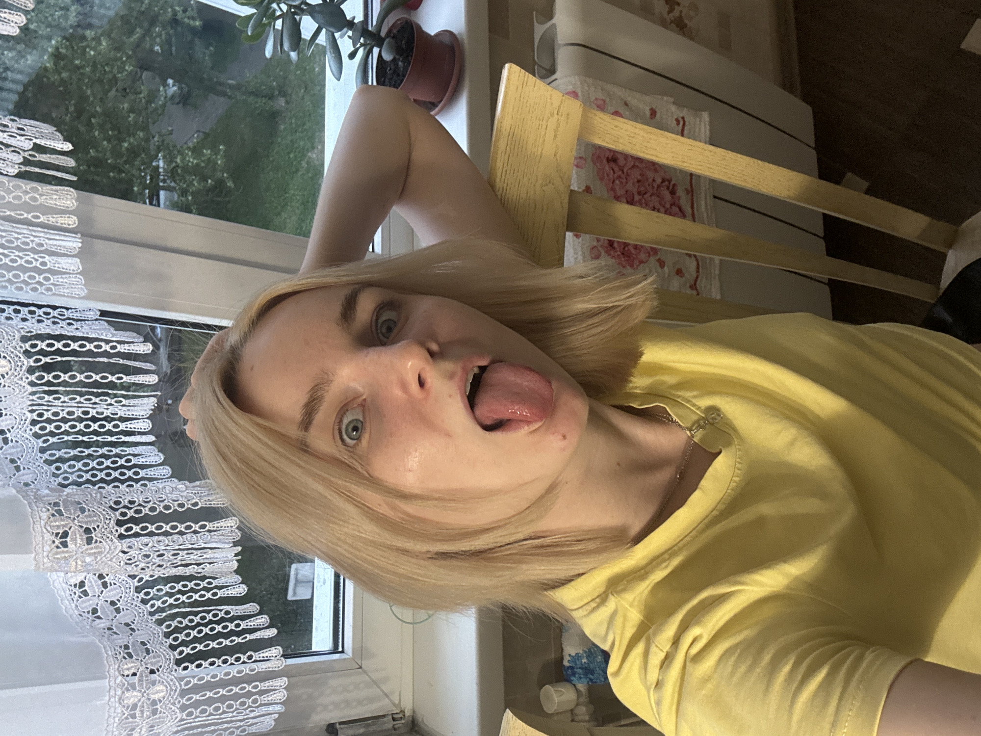 A young woman with blonde hair, wearing a yellow t-shirt, is taking a selfie. She is leaning back in a chair with her right arm behind her head. Her tongue is sticking out. She is positioned in front of a window with white lace curtains. A potted plant is visible on the windowsill. The background outside the window appears to be a green, tree-filled area. The image suggests an indoor setting, likely a residential room. The specific location in the city of Sinyavino, Russia is not determinable from the visual information provided.