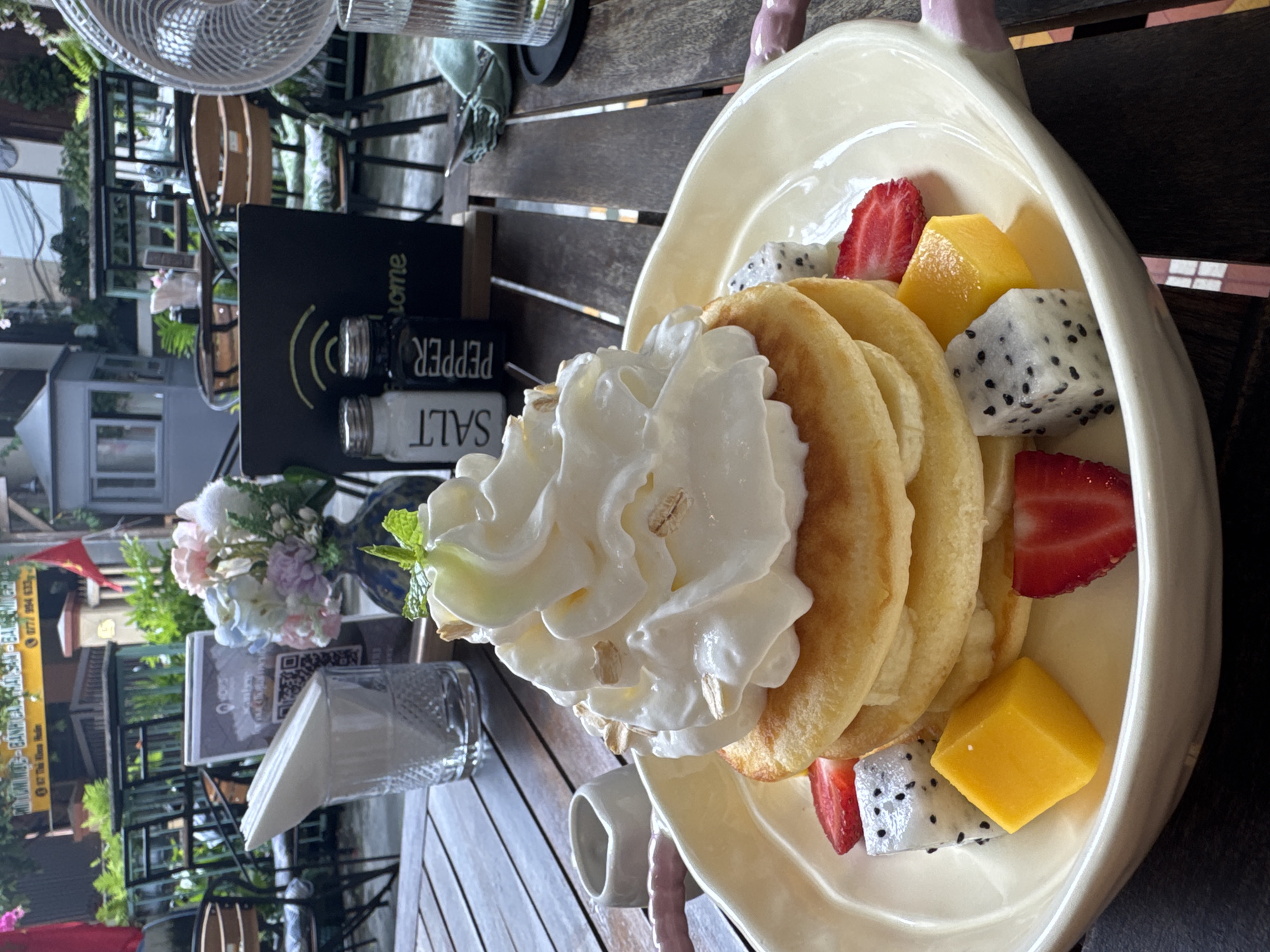 The image depicts a plate of pancakes with whipped cream, strawberries, mango, and dragon fruit, likely photographed at a cafe in Đà Nẵng, Vietnam. The pancakes are stacked and topped with a generous portion of whipped cream and some oats. Sliced strawberries, cubed mango, and dragon fruit segments are arranged around the pancakes. The plate is creamy-colored and has decorative handles. The background shows a wooden table on a terrace or outdoor seating area. There is a fan and salt and pepper shakers on the table. Also, a vase of flowers and promotional material are visible on the table. Through the terrace's edge, part of the surrounding street can be seen.