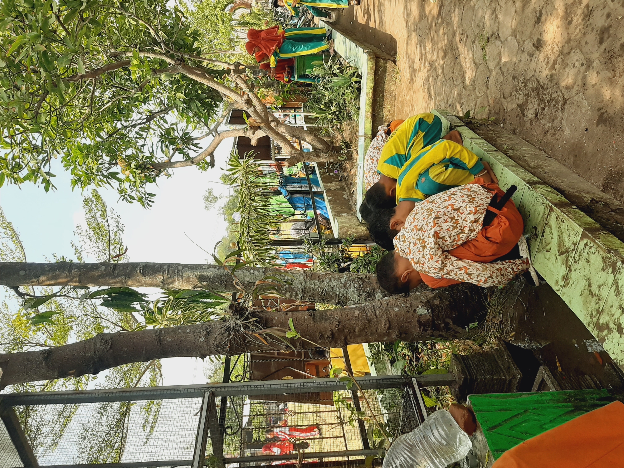The image depicts an outdoor scene in the city of Sumberjo, Indonesia, with several children engaged in activities. In the foreground, three children are crouched, appearing to interact with a water source. One child, wearing an orange outfit and a patterned shirt, is leaning forward. Another child, in a yellow and green uniform, is also leaning down. A third child, also in a yellow and green uniform, is partially visible. To the left, a plastic water bottle is being held by an unseen hand over a green structure. In the background, behind a metal fence, a group of children in red and white uniforms and others in blue and green uniforms are visible. One child in the background is wearing a red hijab and a green and yellow uniform. The environment includes trees with lush green foliage, a stone-paved ground, and a raised concrete border. The overall setting appears to be a school or public area.