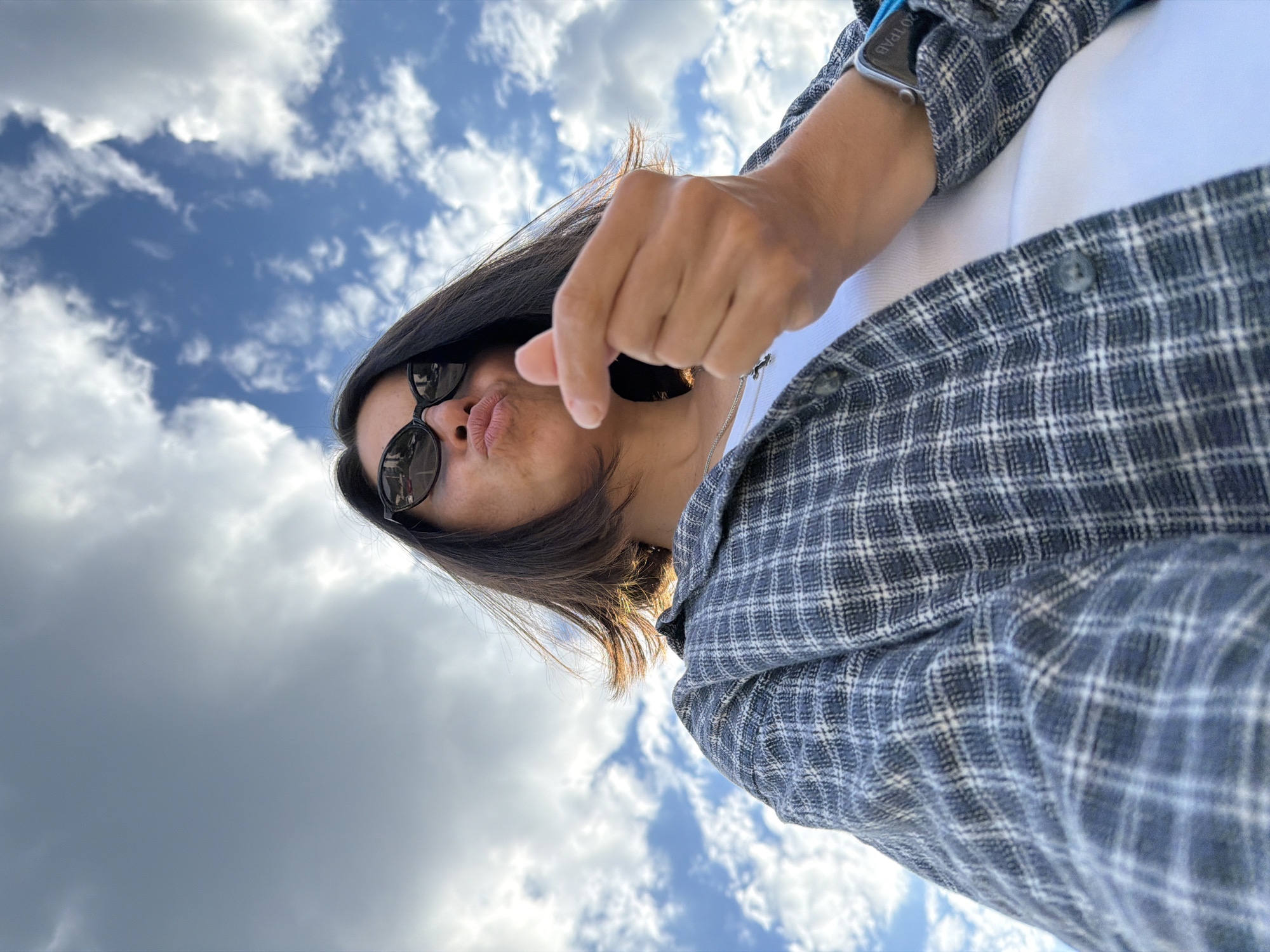 A low-angle view depicts a person outdoors. The person has dark hair and wears sunglasses and a checkered shirt over a white top. The person's lips are puckered in a kissing gesture, and they are holding their hand up towards the camera. The background shows a partly cloudy sky. There is no information about the location being in Budva, Montenegro.