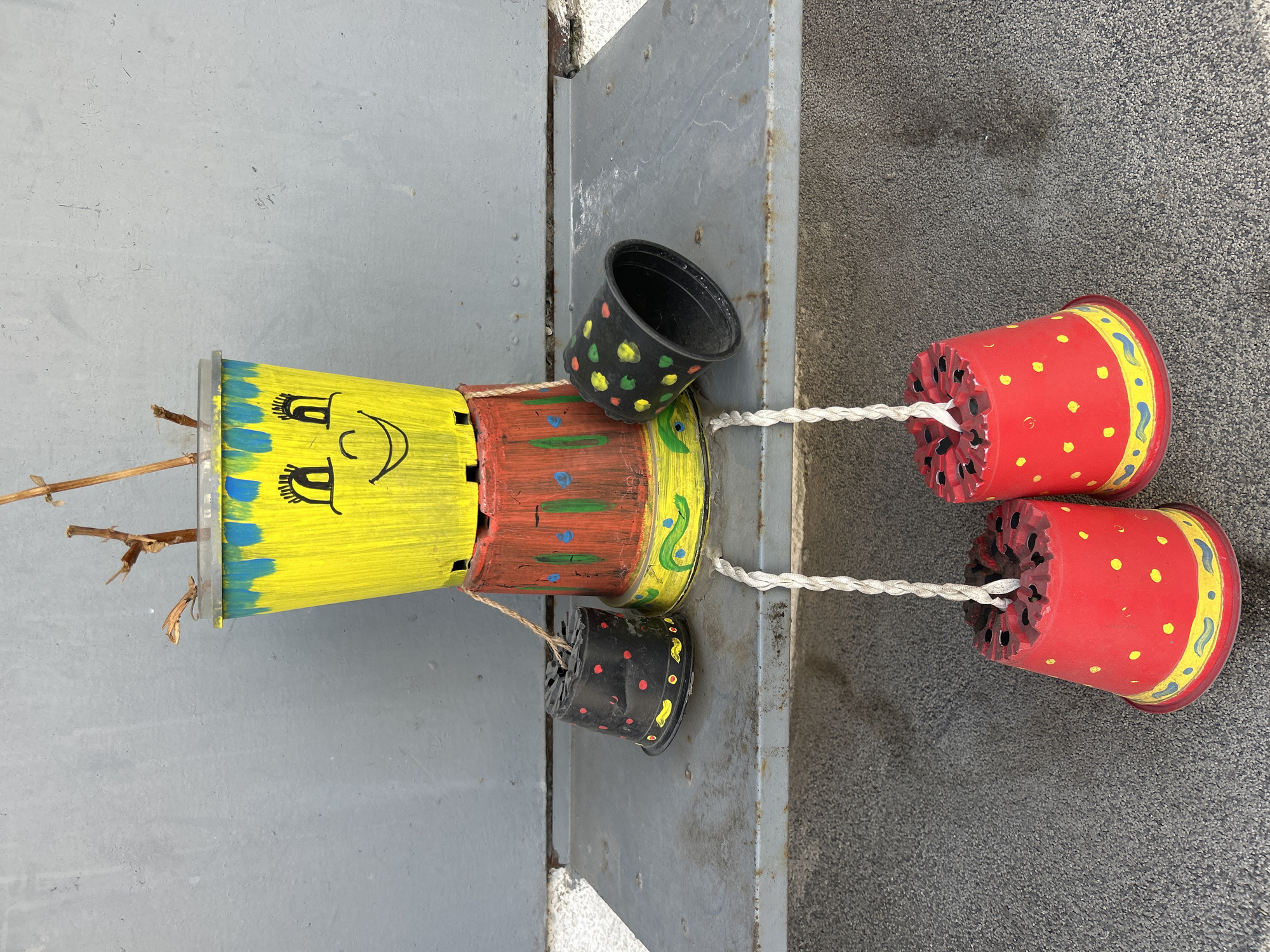 The image shows a handmade doll lying on an outdoor surface that appears to be a metal or concrete platform. The doll is constructed from painted flower pots of various sizes and colors. The doll’s head is yellow with a drawn-on face in black marker, featuring eyes and a smiling mouth. Its torso is reddish-brown. The doll has smaller black pots on either side, acting as hands, and larger red pots for feet. White strings connect the feet and hands to the body. Dry twigs protrude from the top of the head. The background is a mix of gray surfaces, with the texture suggesting different materials. The doll is outdoors and appears to be resting on an edge or ledge. There is no indication of a specific location within Plovdiv, Bulgaria.