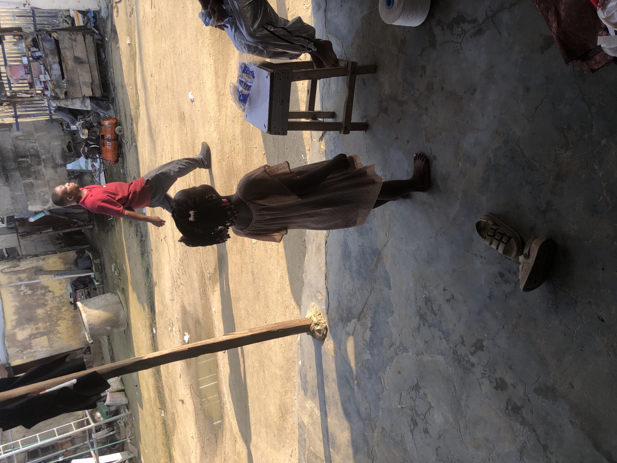 This image captures a scene in Ado, Nigeria, likely an outdoor marketplace or workshop area. In the foreground, a young girl with braided hair, wearing a light-colored dress, stands barefoot on a cracked concrete floor. She is facing away from the camera, looking towards the right. To her right, a simple wooden stool holds several items, including what appears to be packaged goods and paper. A seated person, partially visible on the far right, wears silver-colored pants. A large spool of white thread is also present.
In the background, a man in a red polo shirt and grey trousers walks away from the camera. Behind him, an orange air compressor is visible. The environment is dusty with a few puddles of water on the ground. Various structures, including buildings with weathered walls and metal fencing, suggest a busy and perhaps informal commercial or industrial zone. A ladder leans against a structure on the left. A wooden pole, possibly part of a temporary shelter, is prominent in the middle ground. A pair of sandals lies on the concrete floor in the lower left portion of the image.